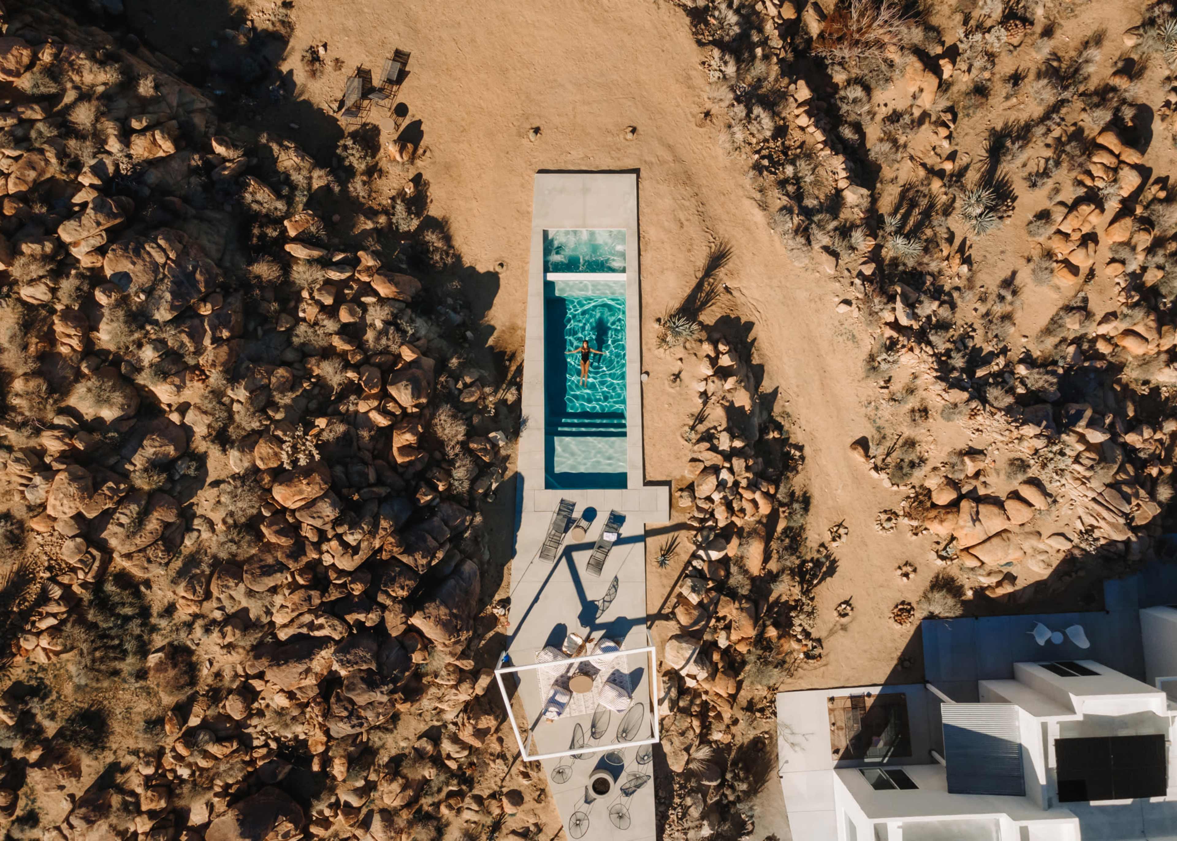 An aerial view shows a swimming pool surrounded by rocky terrain and a modern building in the lower right corner.