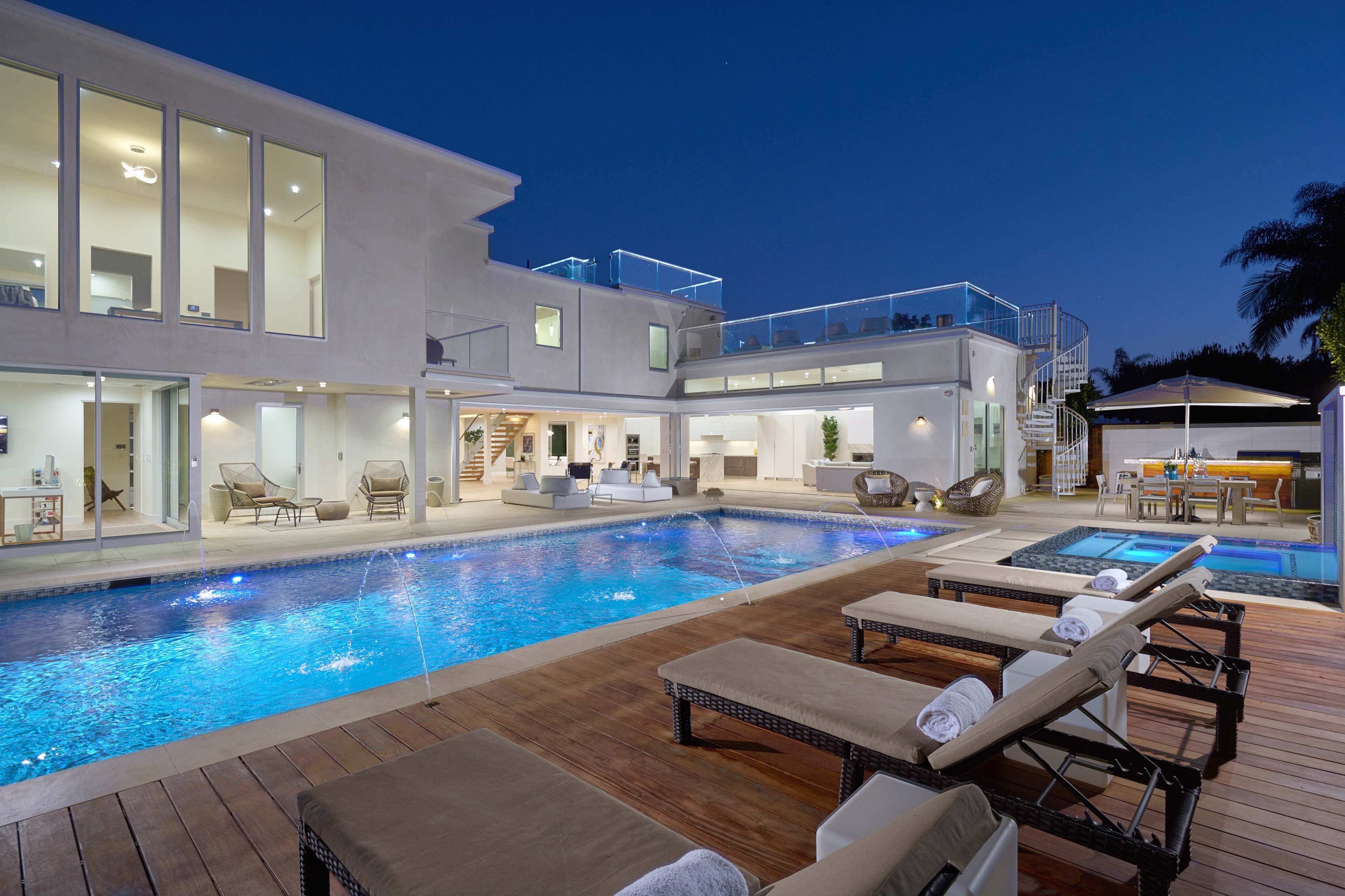 A modern two-story villa with a large swimming pool and lounge chairs, illuminated at dusk with outdoor seating areas and a hot tub.