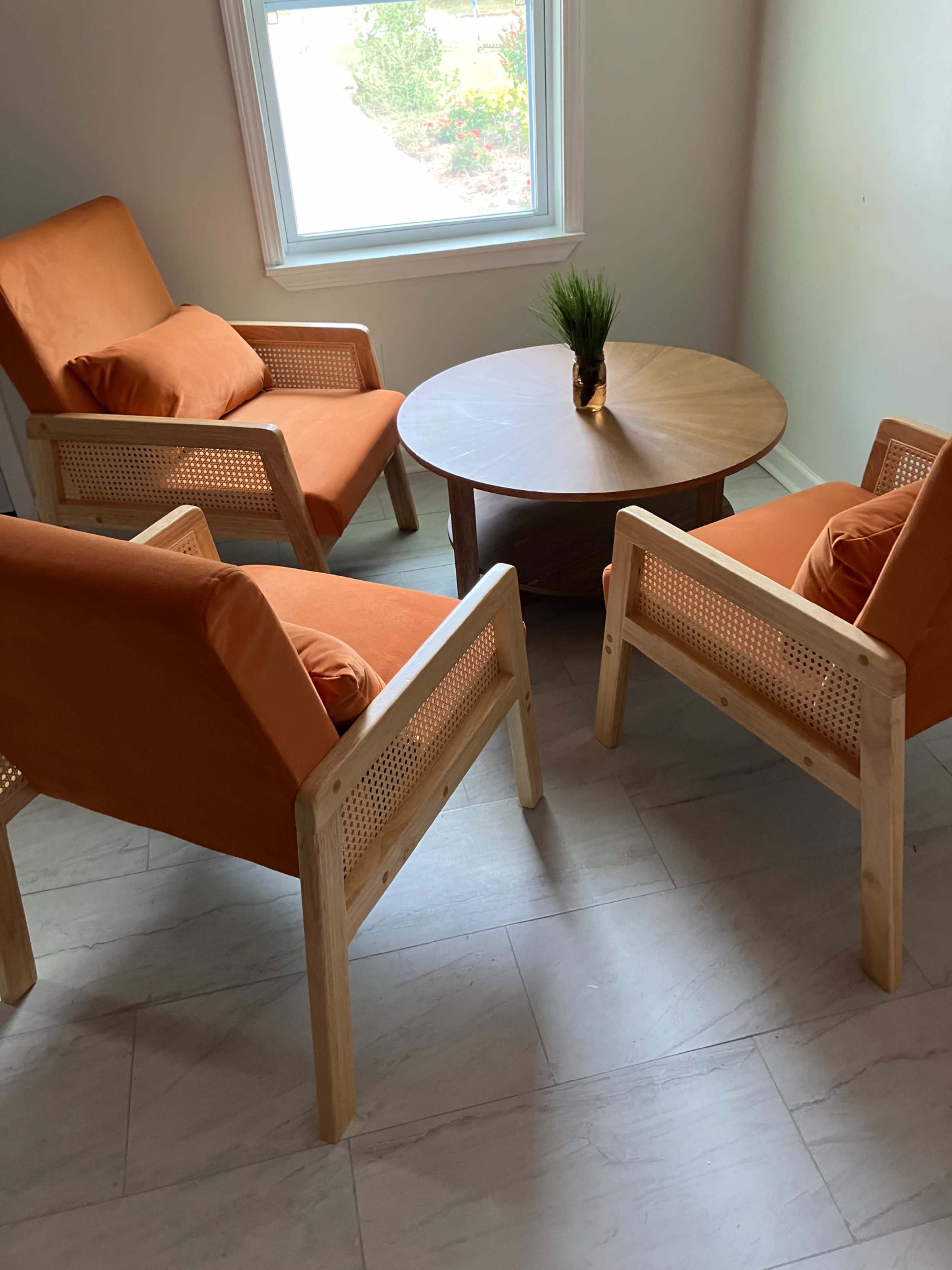 The image shows a small seating area with two orange chairs made of wood positioned around a round wooden coffee table, with a plant in a vase on top of the table, near a window.