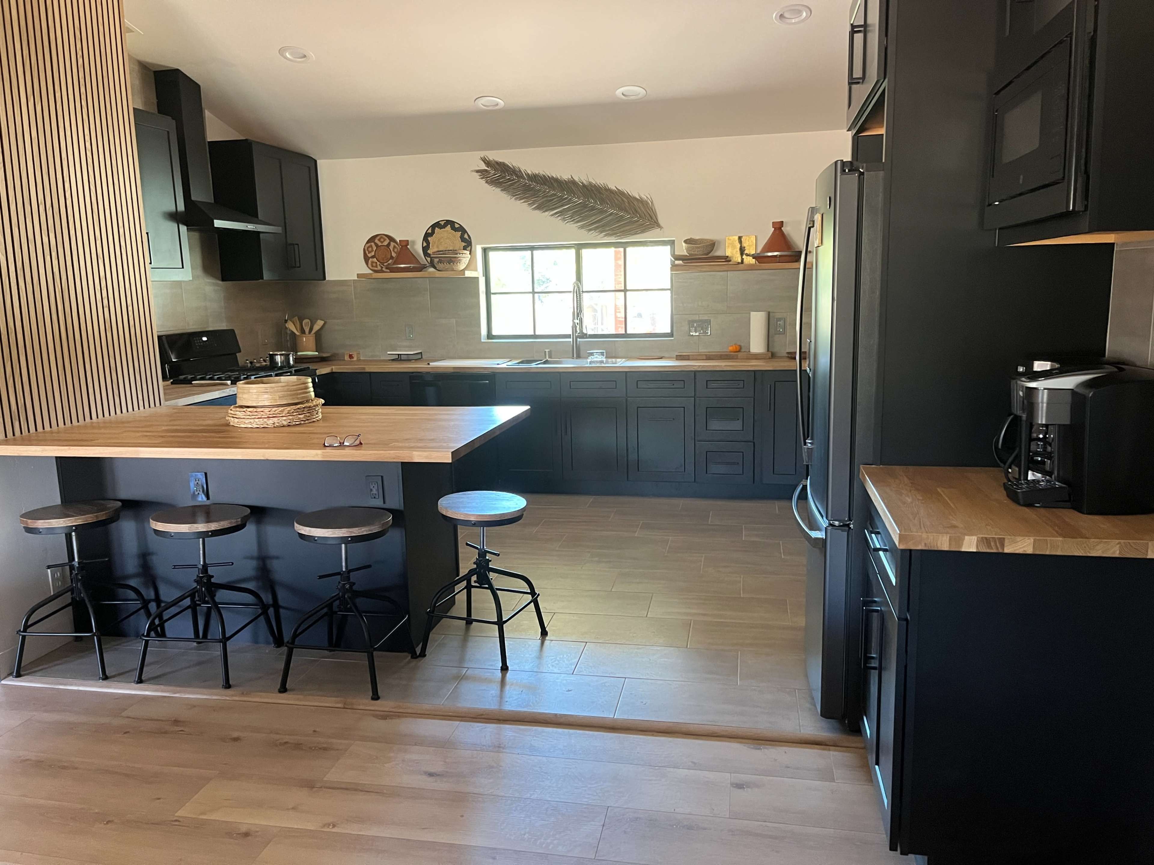 The image shows a modern kitchen featuring black cabinets, a wooden countertop with three stools, and a large window allowing natural light.