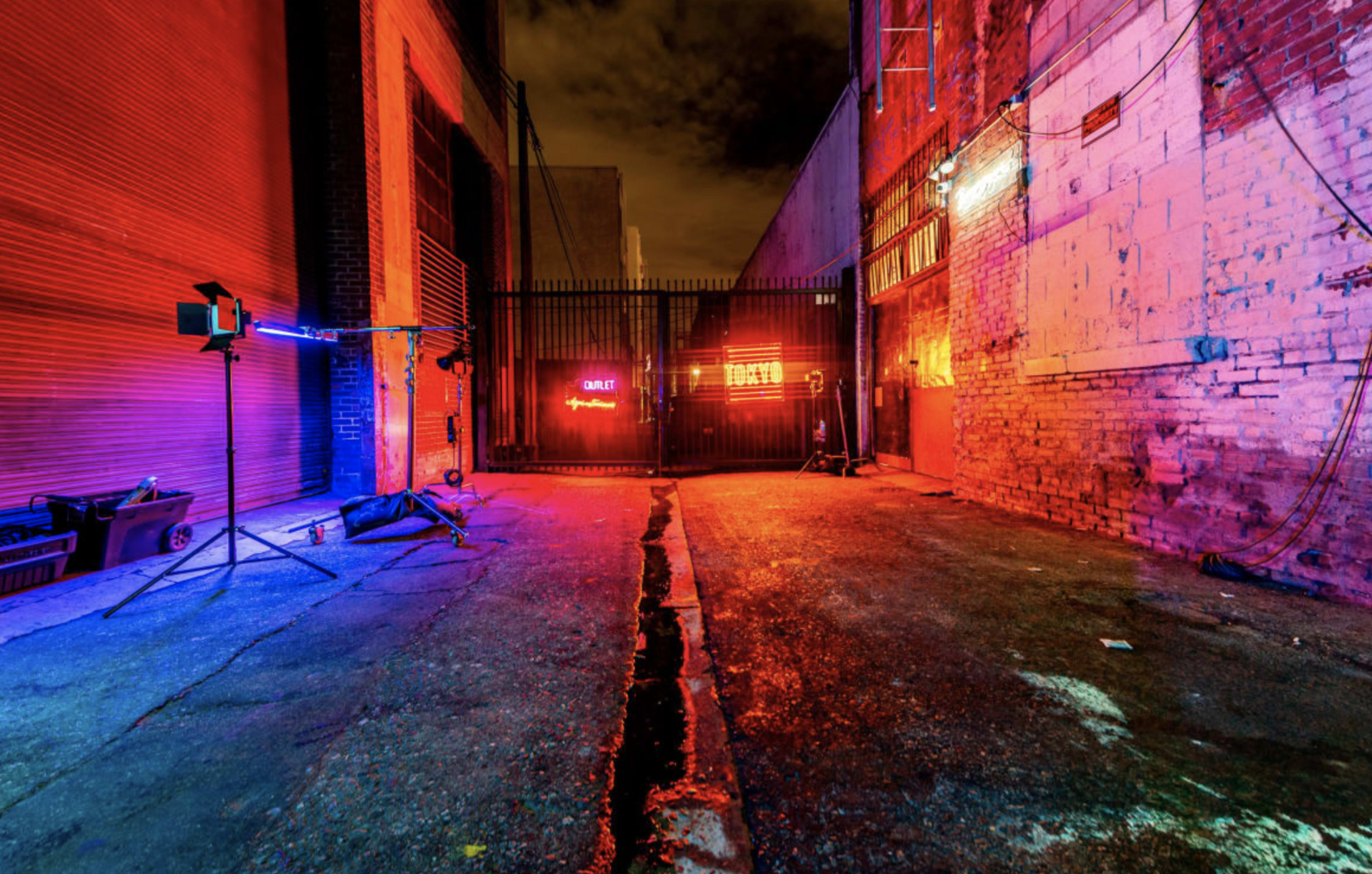 A narrow alleyway features colorful neon lights illuminating the walls and a gated entrance.