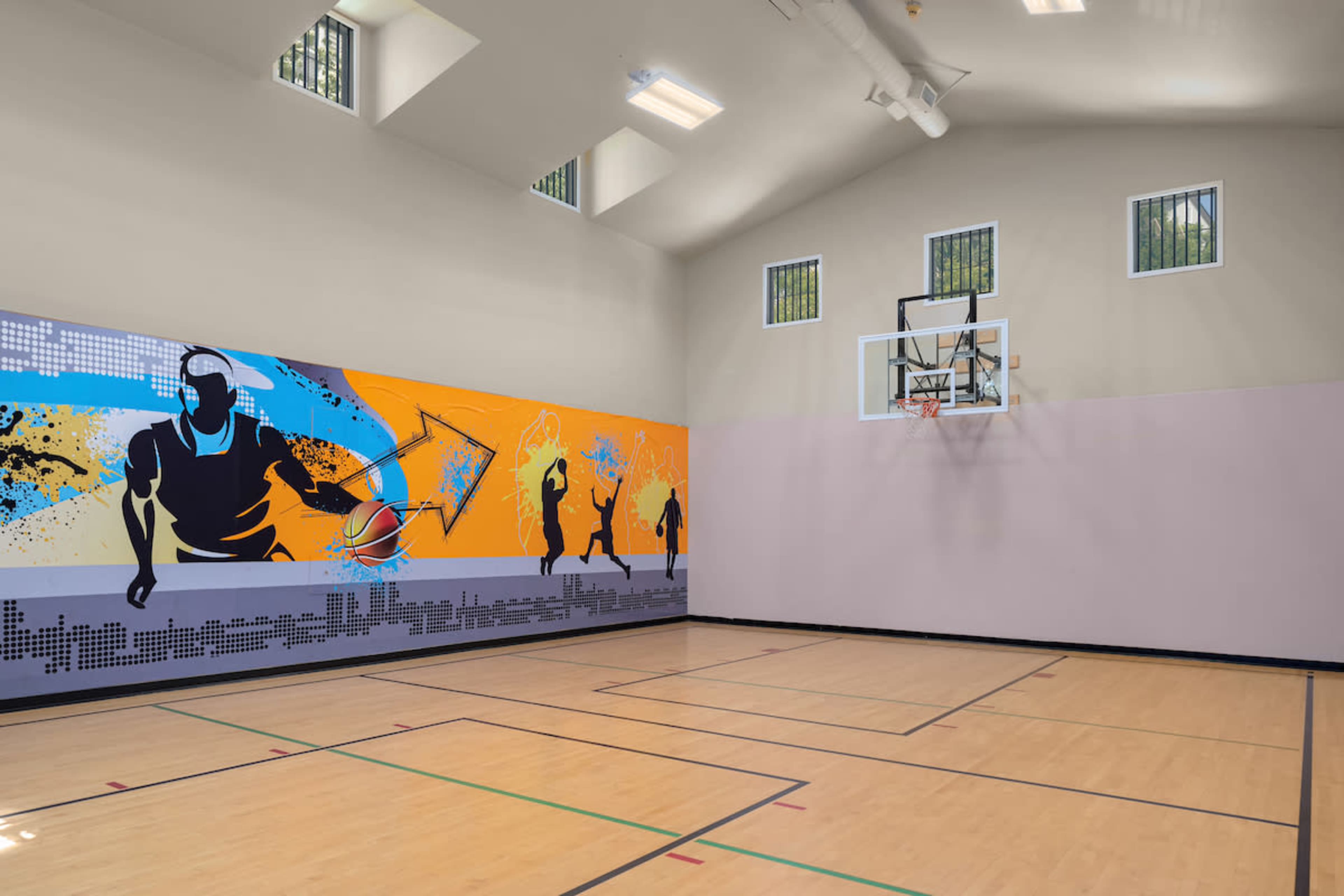 The image shows an indoor basketball court with a colorful mural on one wall and a basketball hoop mounted on the opposite side.