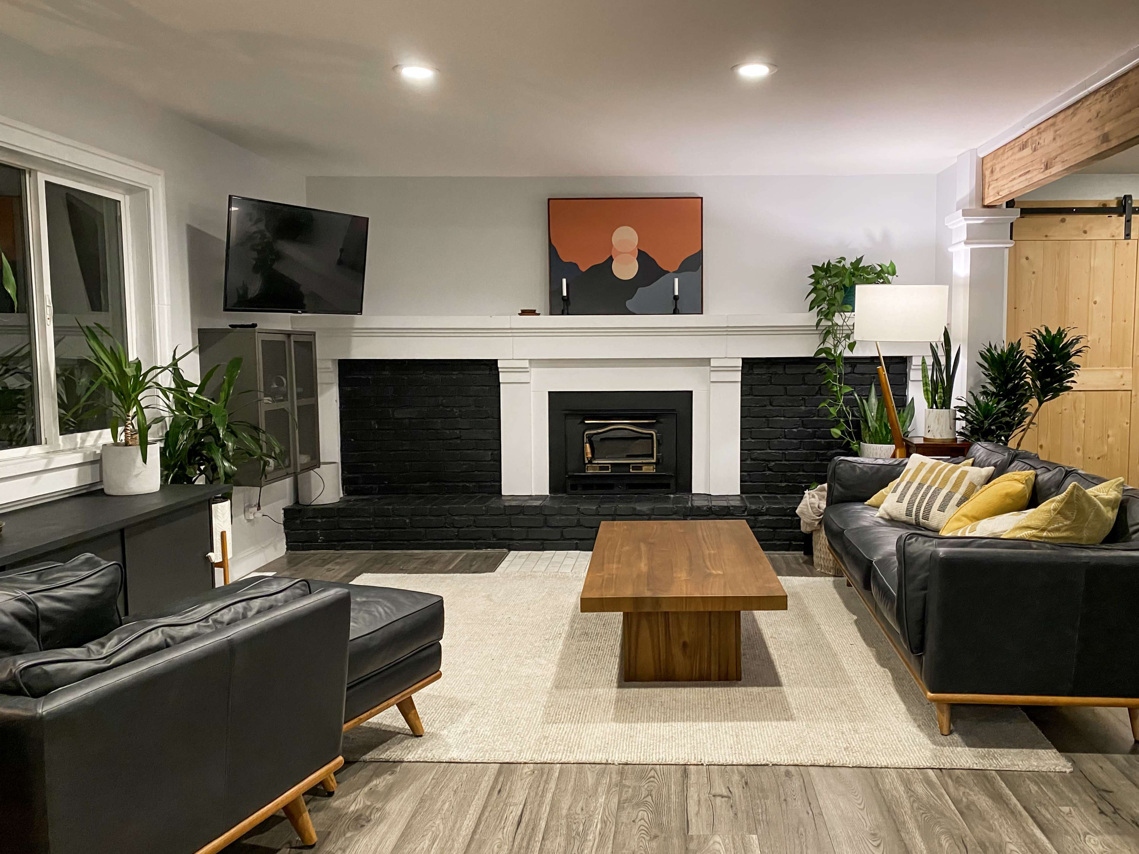 A modern living room featuring two black sofas, a wooden coffee table, a television mounted on the wall, and various plants surrounding a black brick fireplace.