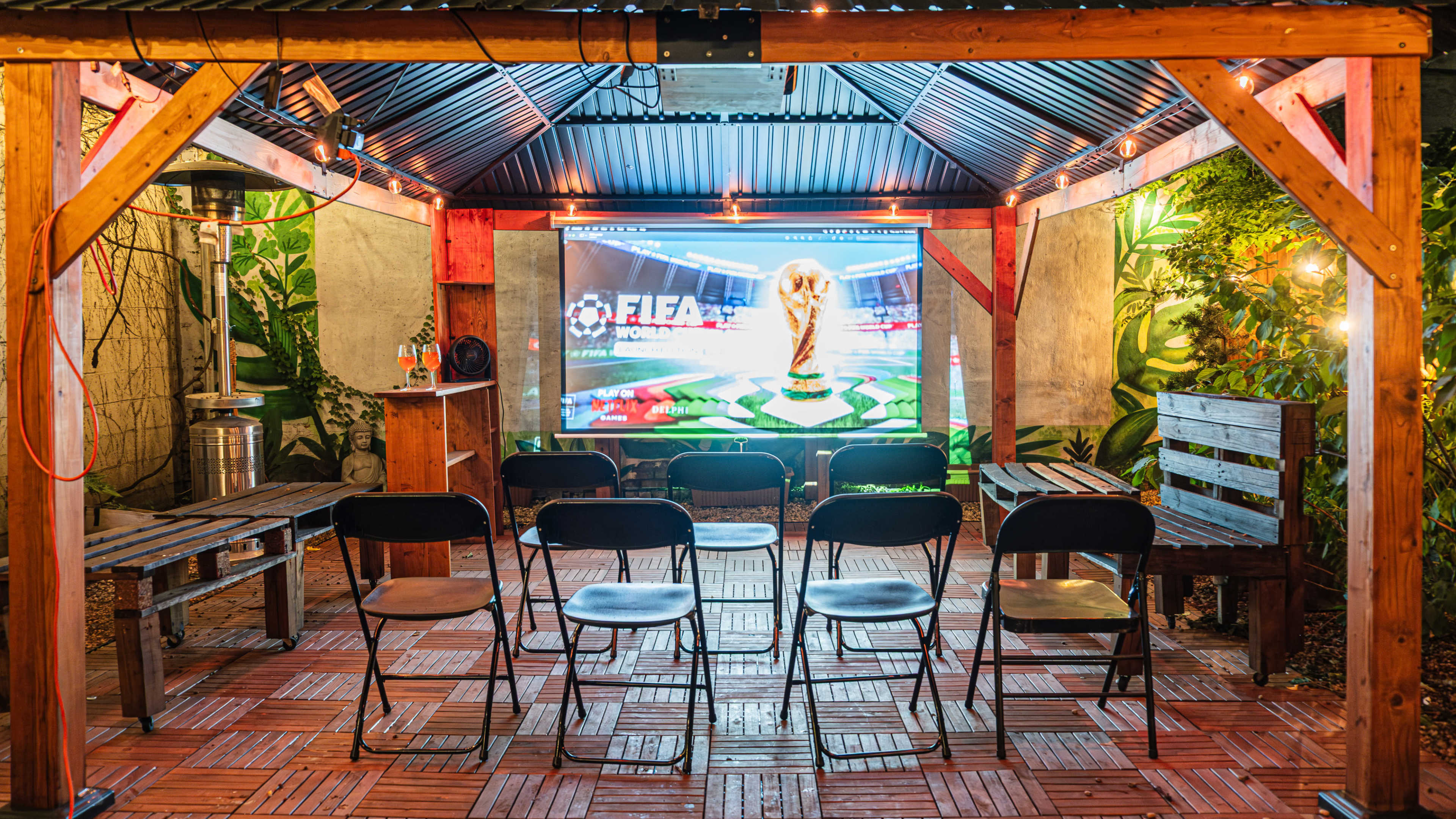 The image shows a semi-outdoor space set up for a viewing event, featuring several black chairs arranged in front of a large screen displaying a FIFA video game.