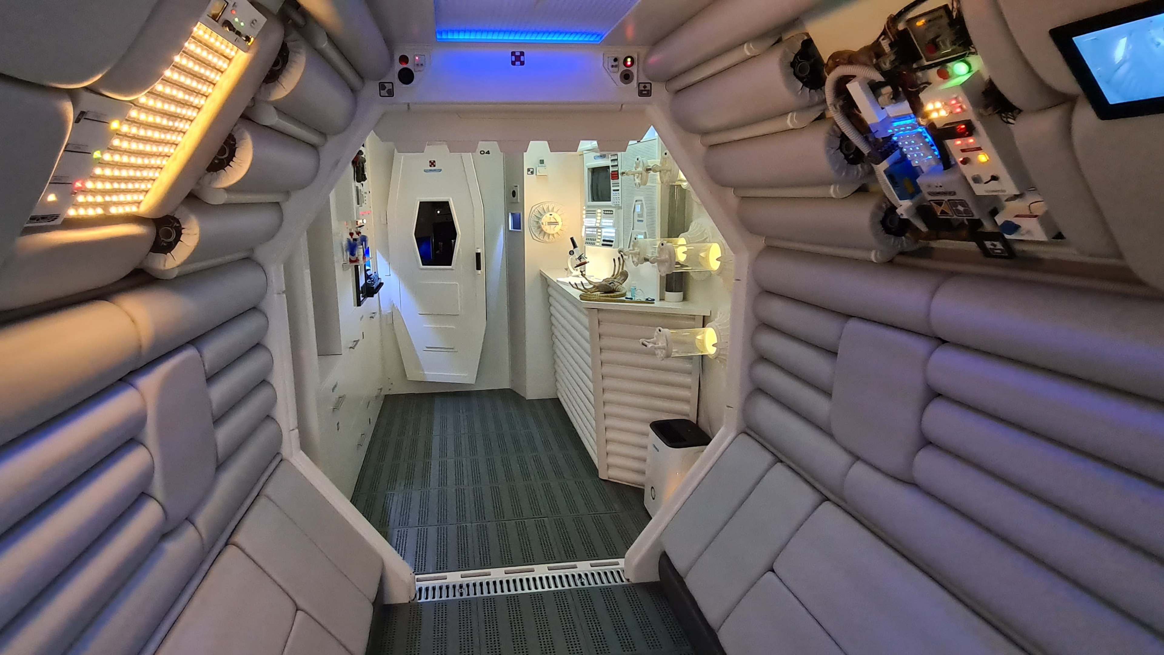 A corridor in a futuristic spacecraft with padded walls, control panels, and doors leading to other sections.