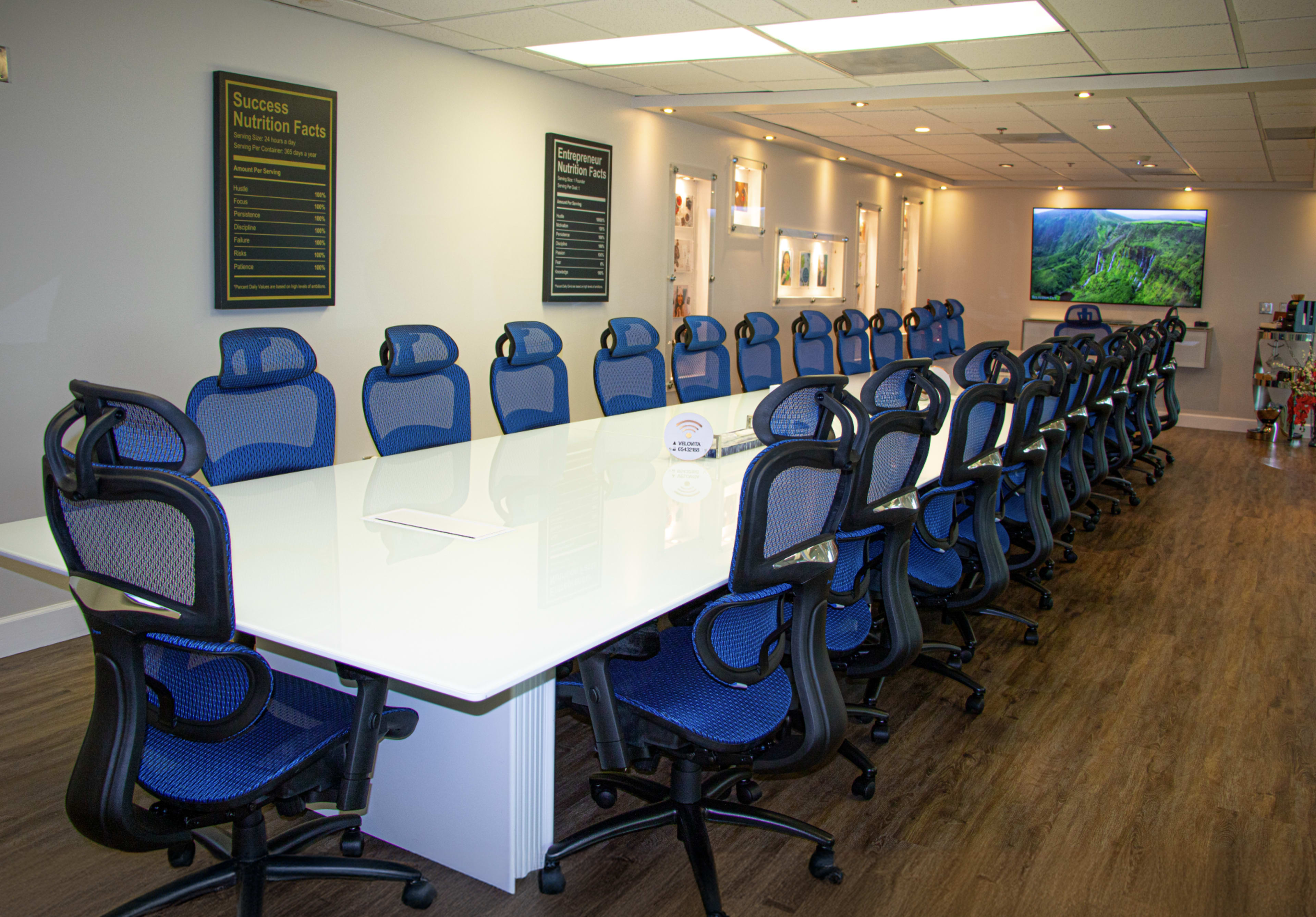 A long, white conference table is surrounded by blue and black ergonomic chairs in a well-lit meeting room.