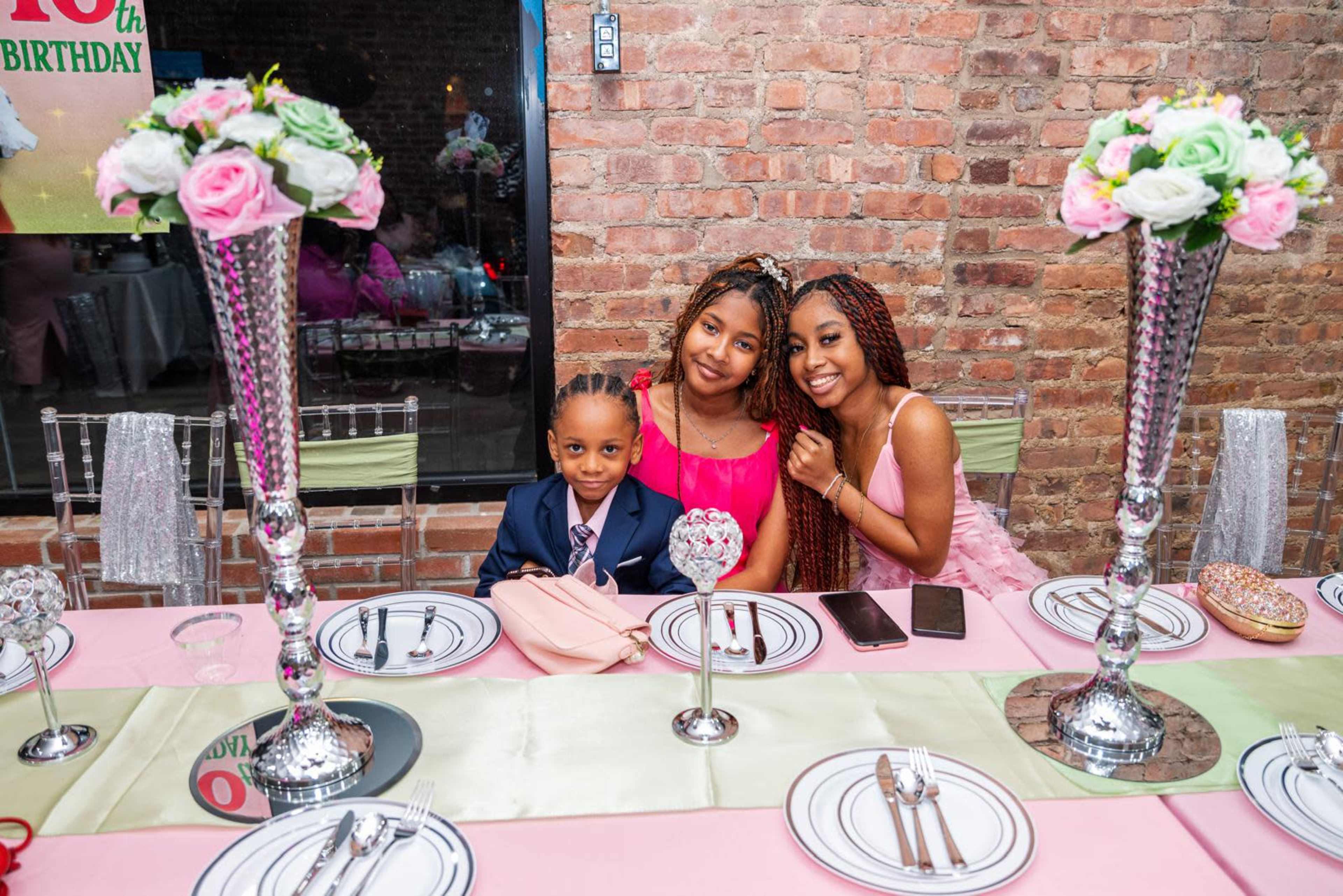 Three children are seated at a festive table decorated with flowers and elegant tableware for a birthday celebration.