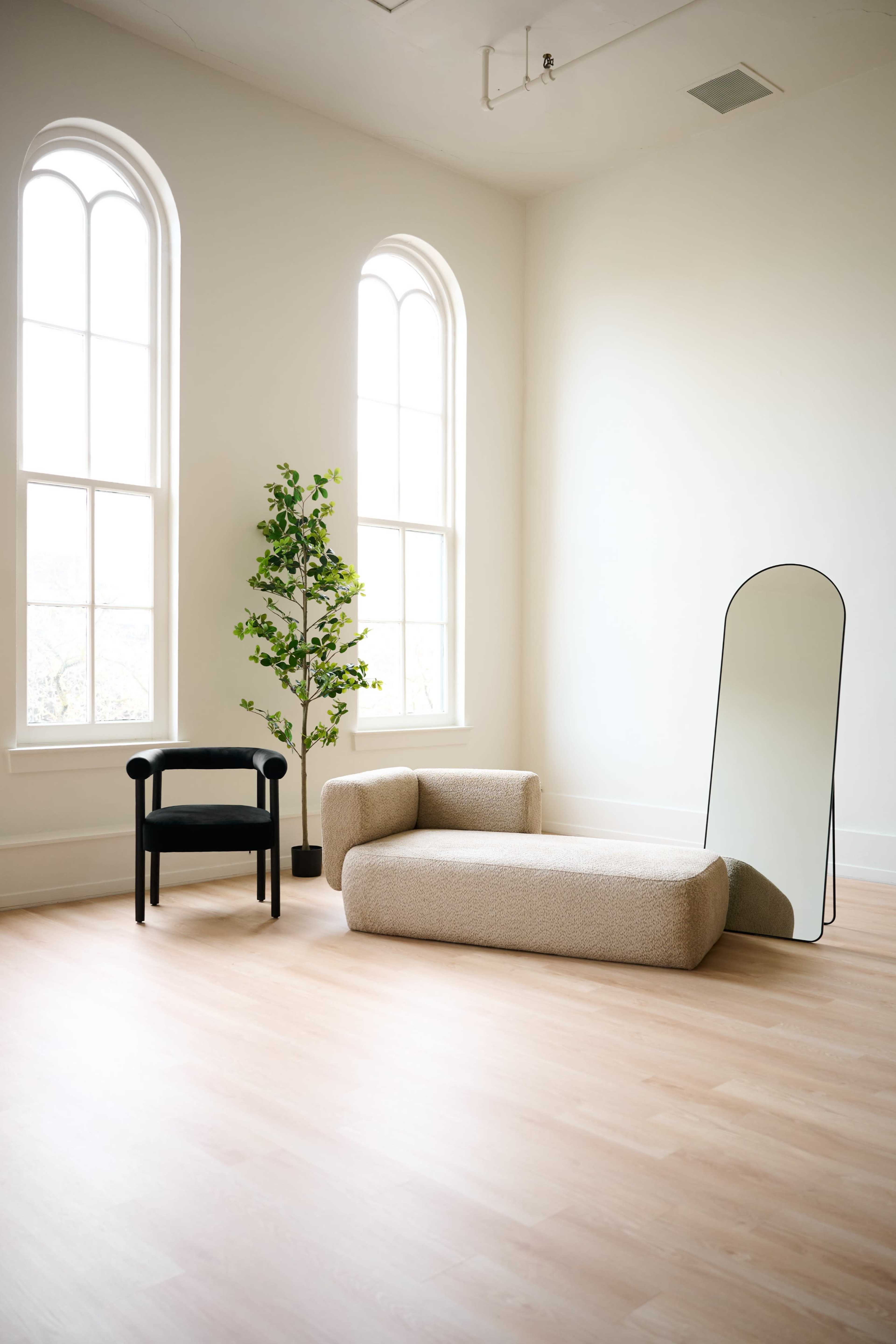 The image shows a minimalist interior with a light wooden floor, a curved beige sofa, a black chair, a tall potted plant, and an arched mirror near large windows.