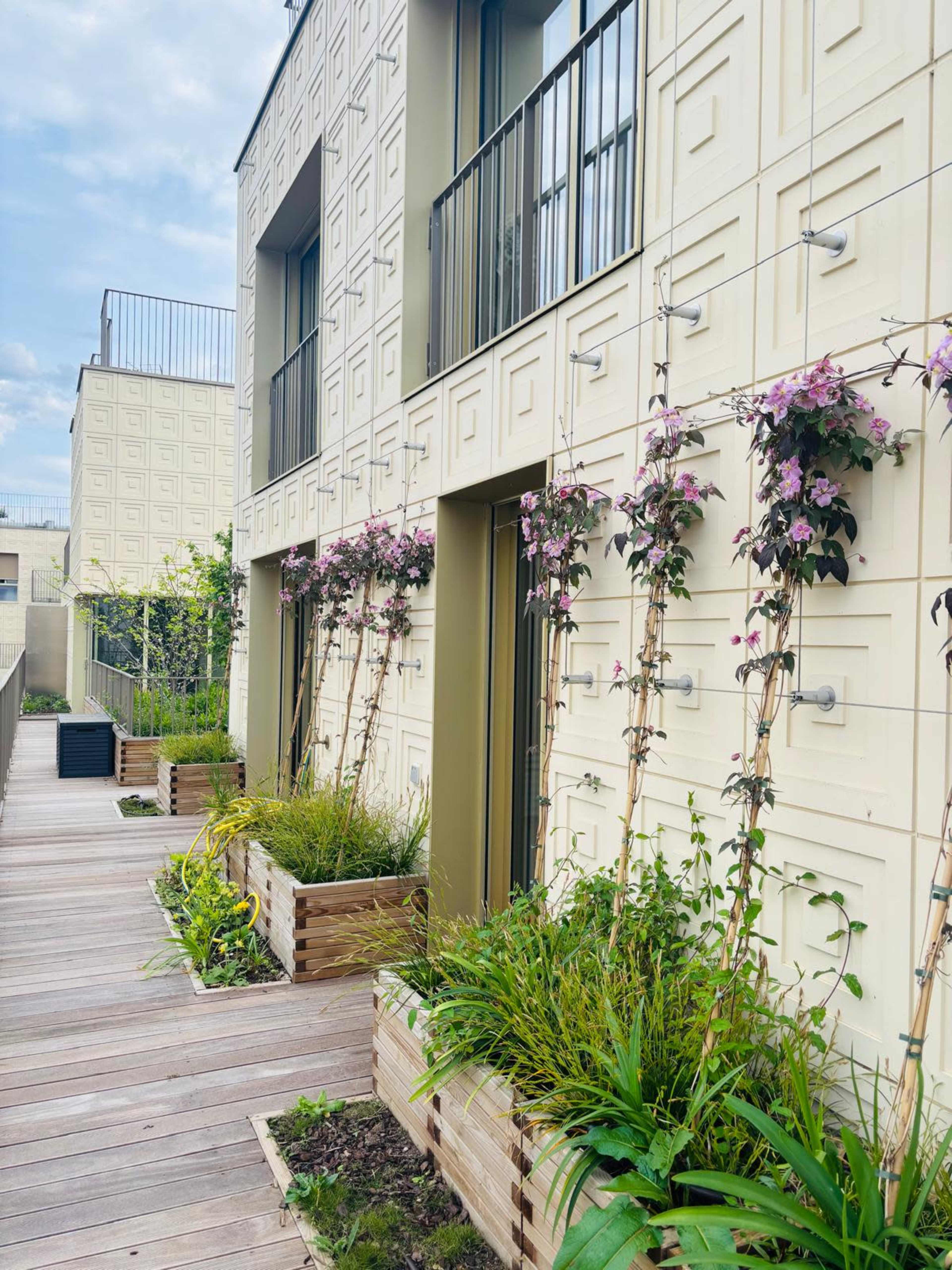 The image shows a modern building facade with geometric patterns, featuring several balconies adorned with flowering plants and greenery along a wooden walkway.