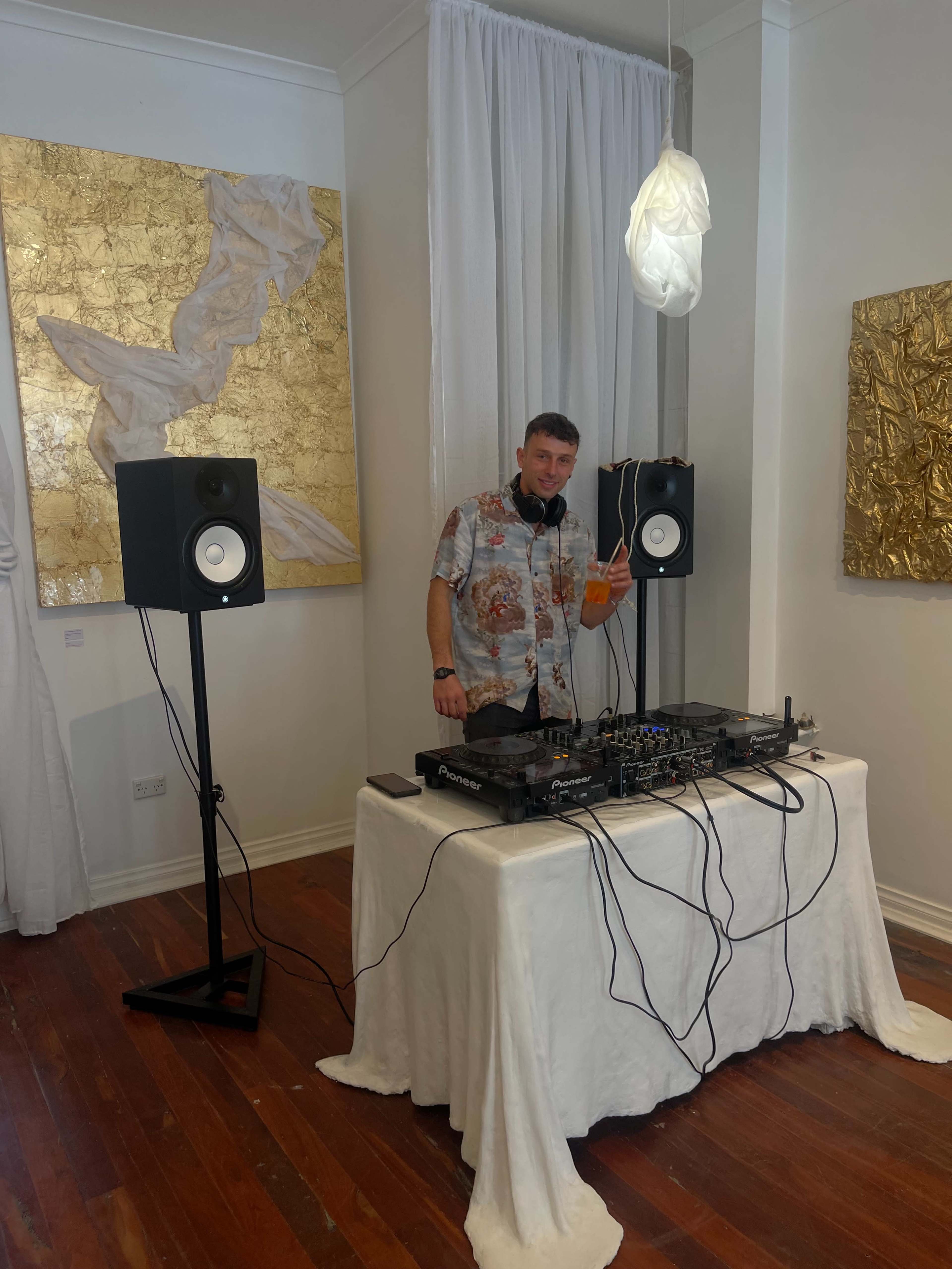 A DJ stands behind a setup featuring turntables and speakers in an art gallery adorned with gold leaf artworks.