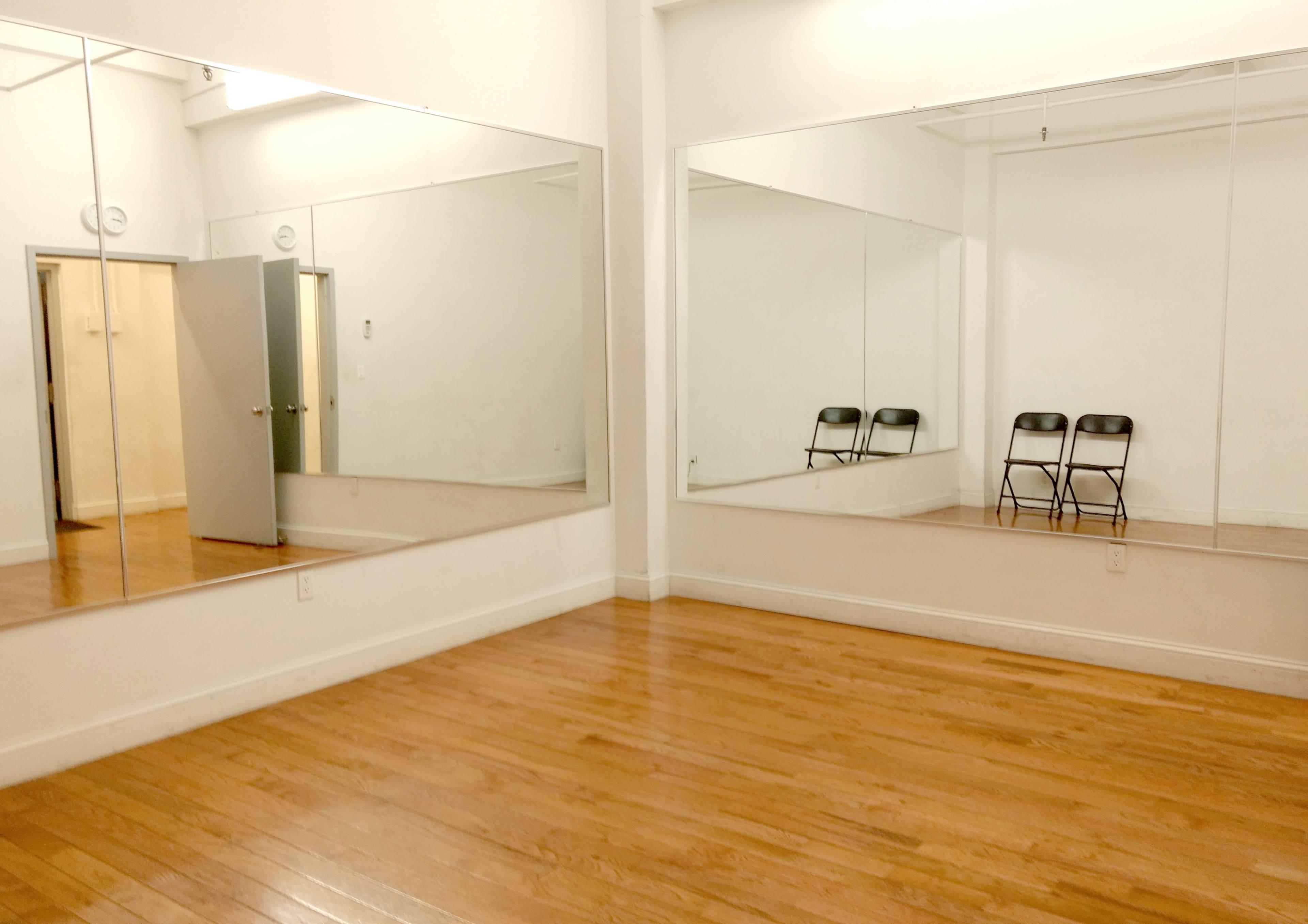 A bright room with wooden floors, featuring two large mirrors on opposite walls and three black folding chairs arranged in front of one mirror.