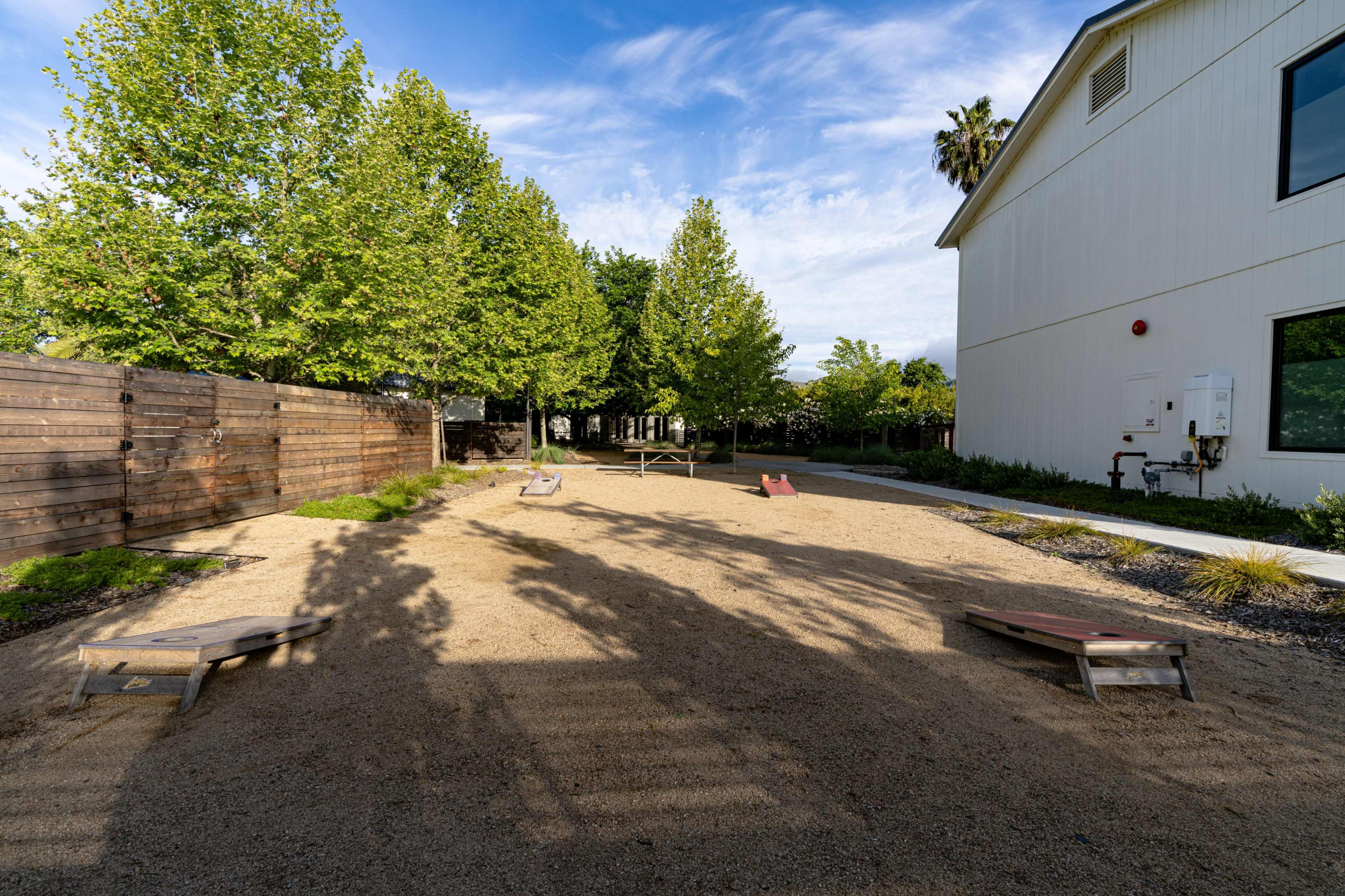 The image shows a gravel area with wooden benches and set up for cornhole games, surrounded by trees and a building.