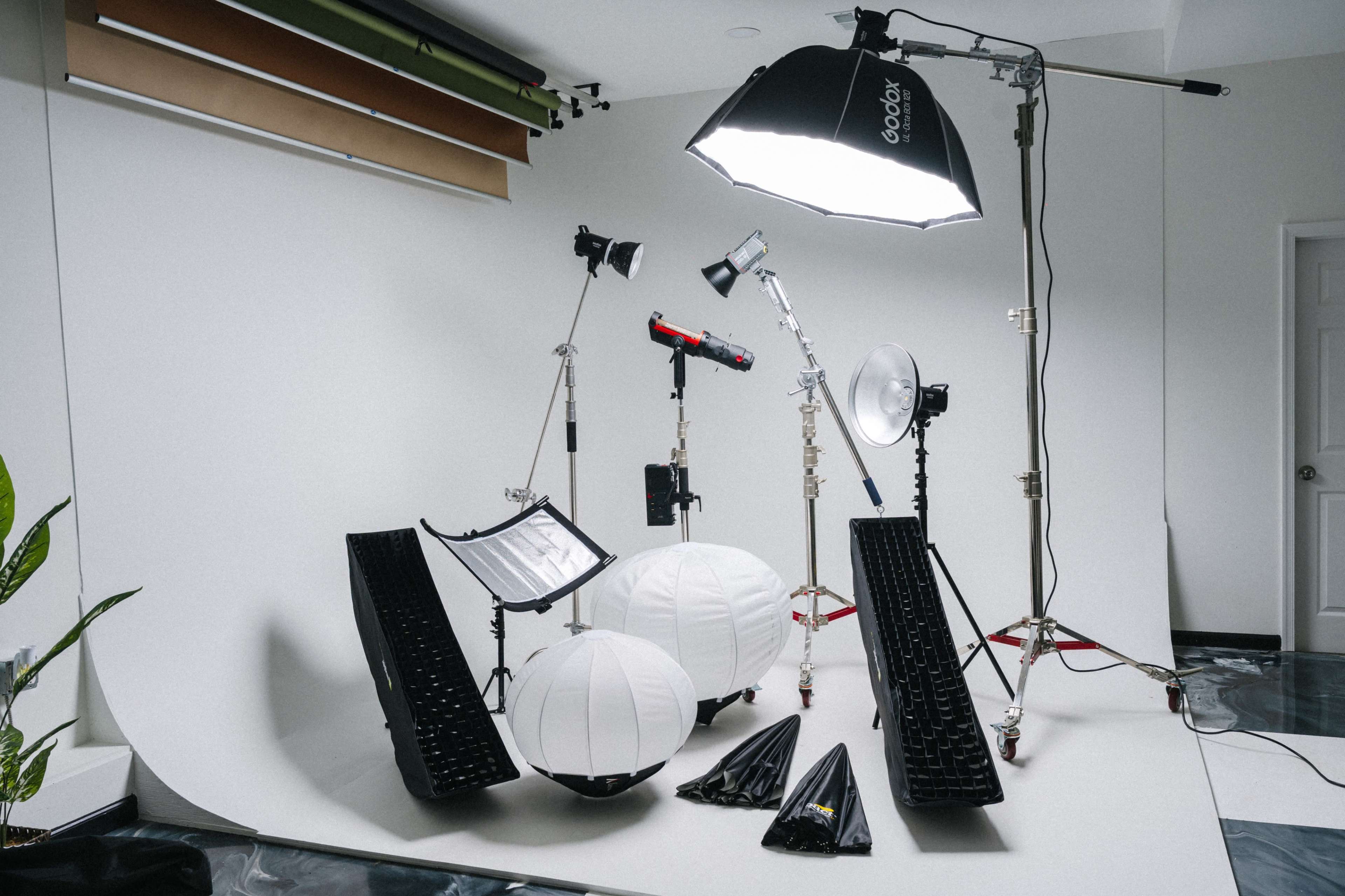 The image shows a photography studio setup featuring various lighting equipment, including softboxes, reflectors, and different light modifiers arranged around a seamless backdrop.