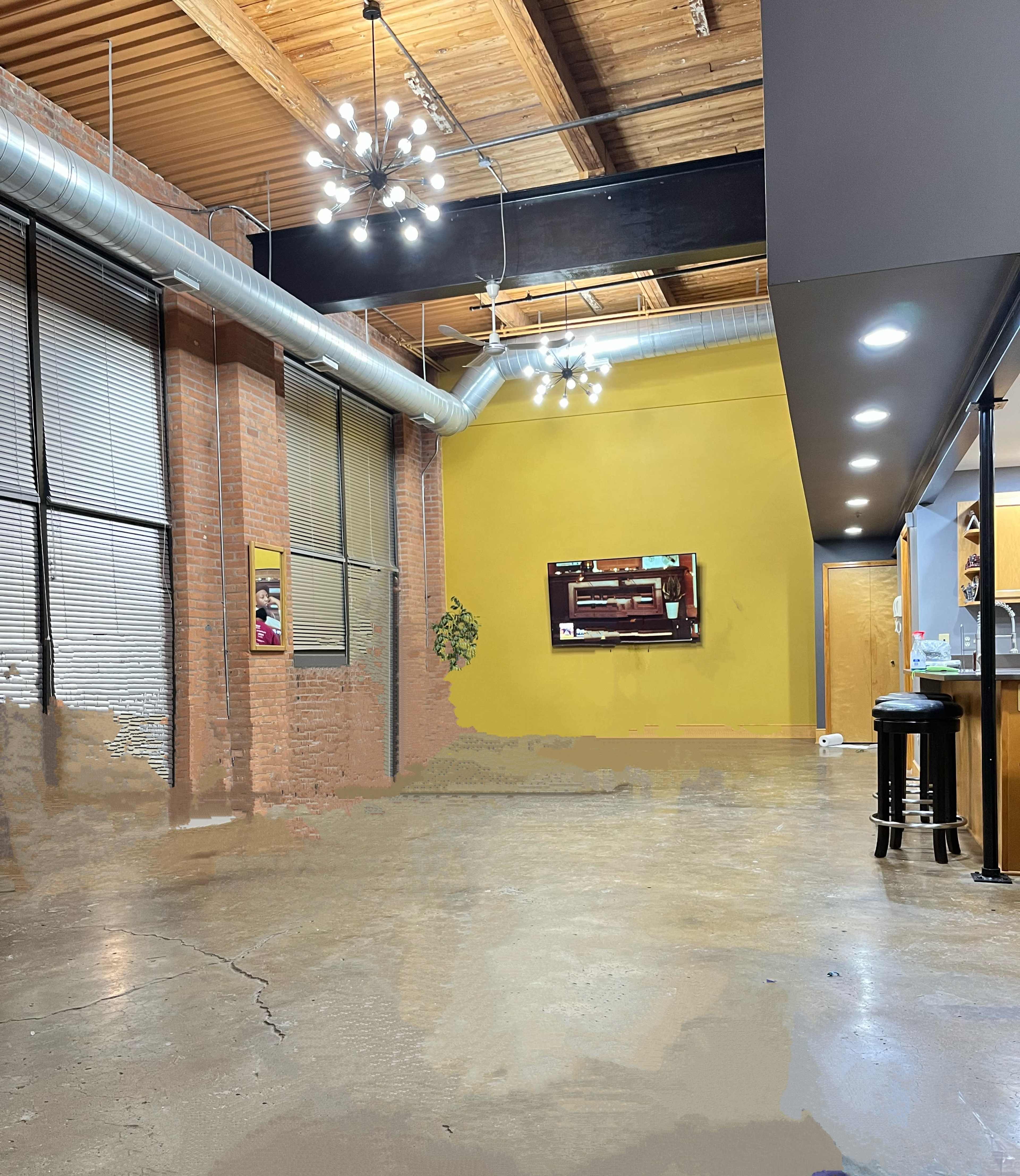 An industrial-style room with a painted yellow wall, exposed brick, and overhead lighting, featuring a bar area and a framed artwork.
