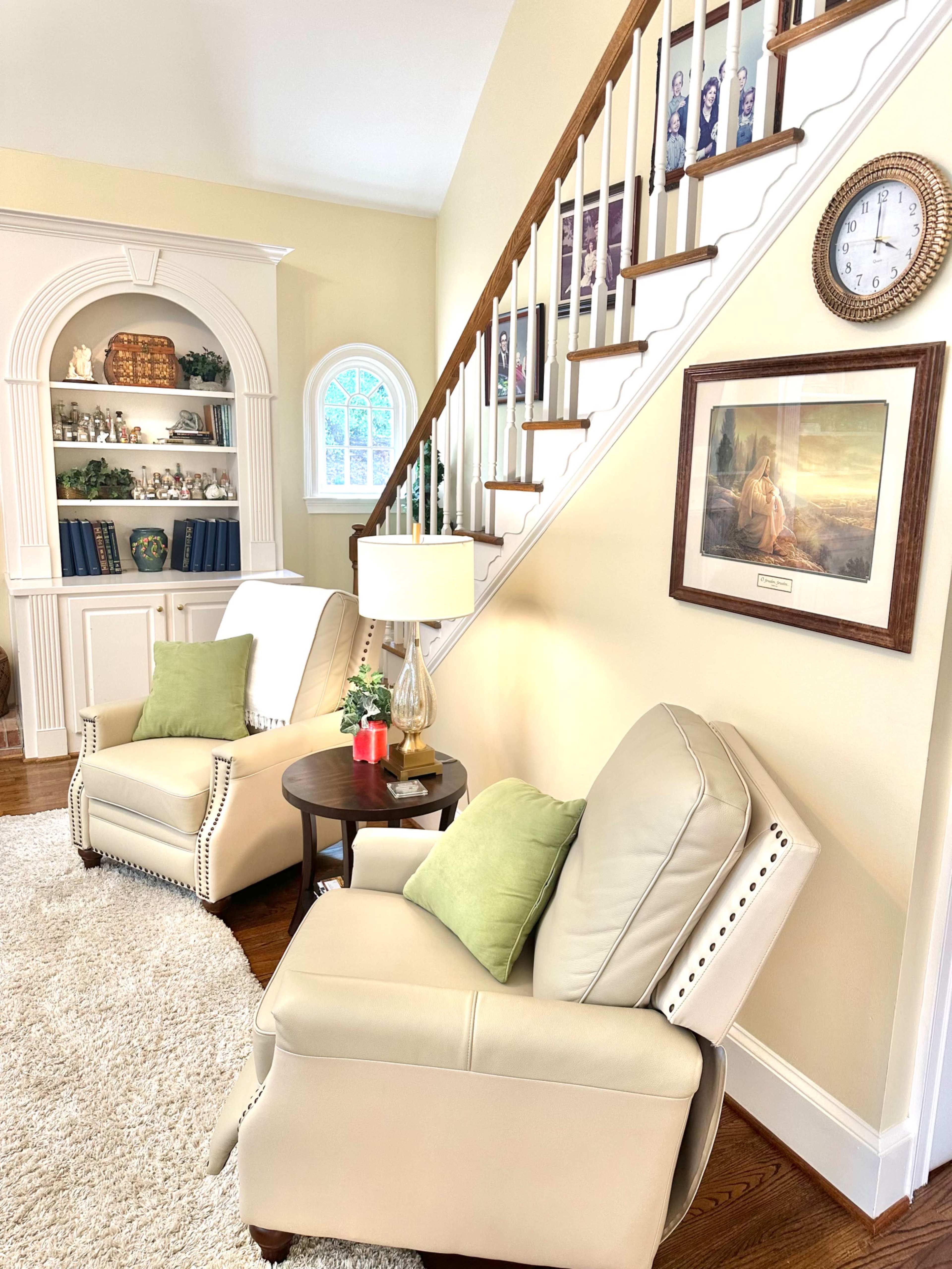 The image features a cozy living room with two reclining chairs, a small side table with a lamp, and a staircase in the background adorned with an archway shelf filled with decorative items.