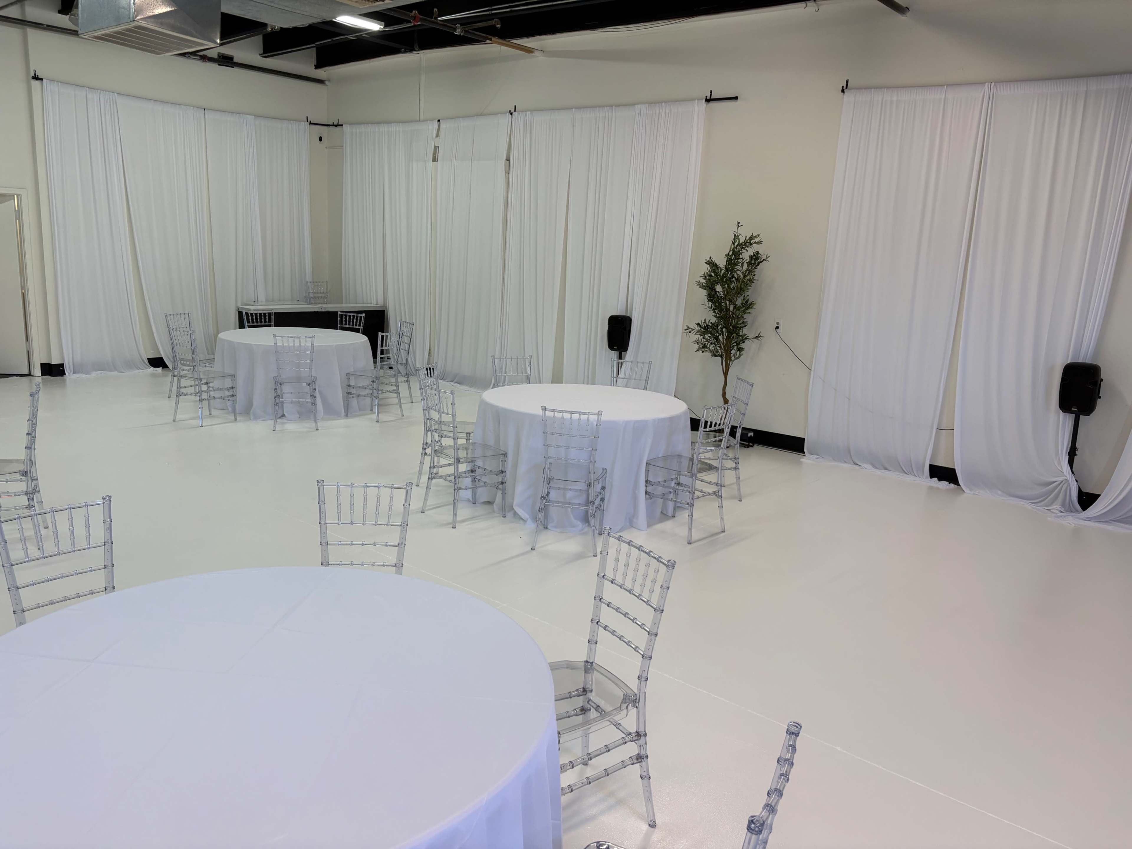 The image shows an empty event space with white walls, white tablecloths on round tables, and transparent chairs arranged around the tables.