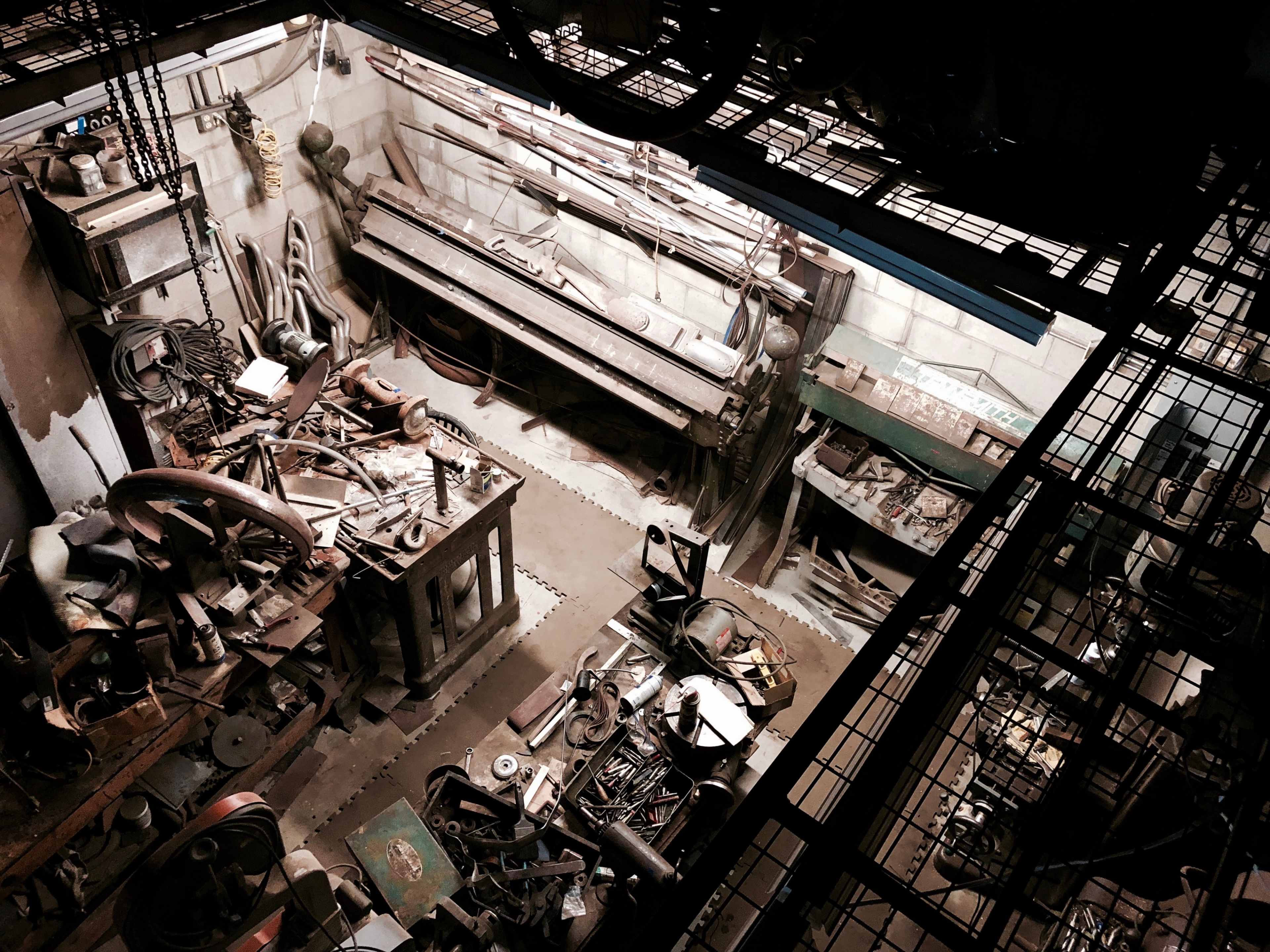 An overhead view of a cluttered industrial workshop filled with tools, machinery, and parts scattered across the floor and workbenches.