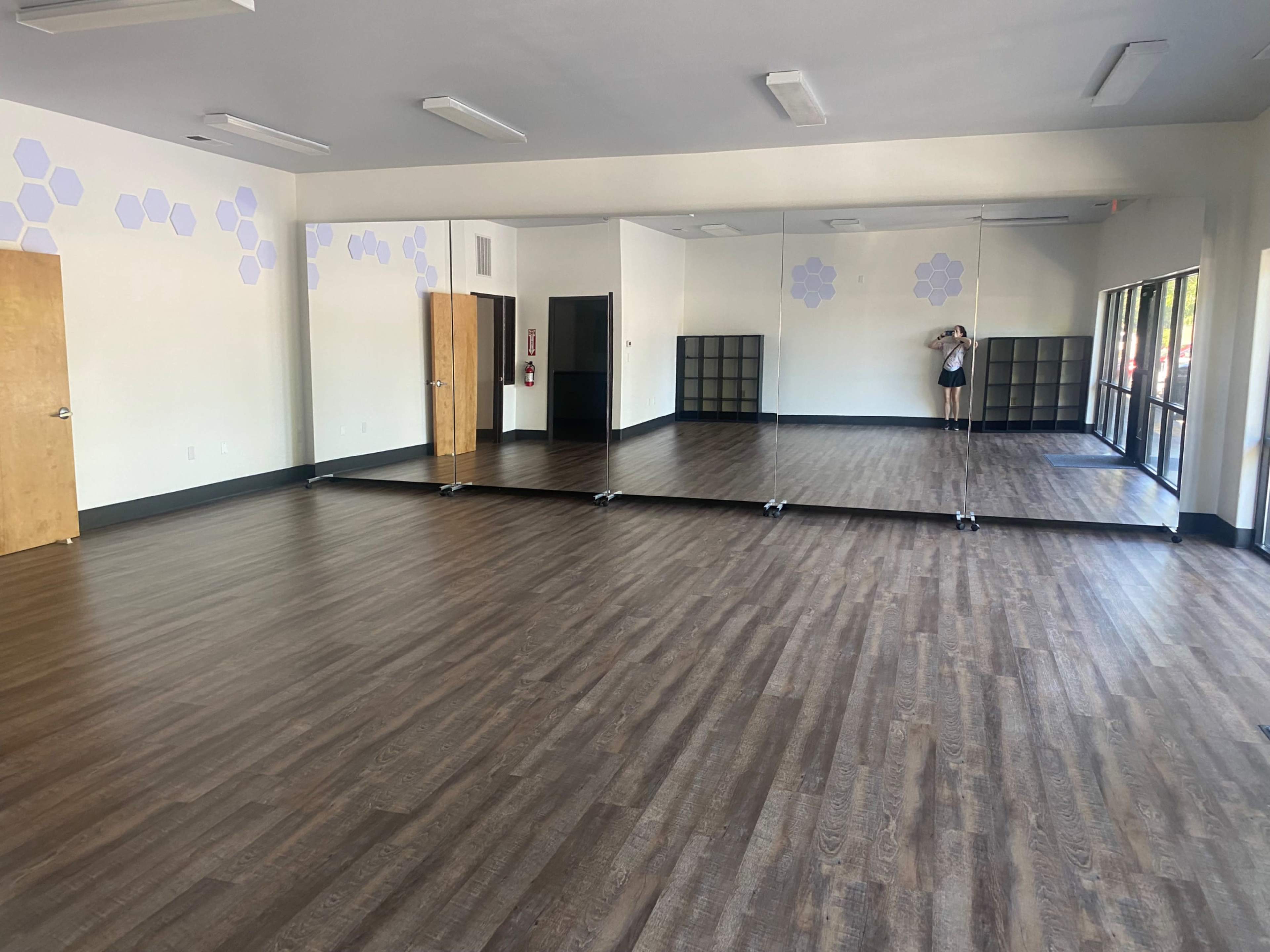 The image shows a spacious, empty room with wooden flooring and a large mirror along one wall, featuring doors and a few shelves in the background.