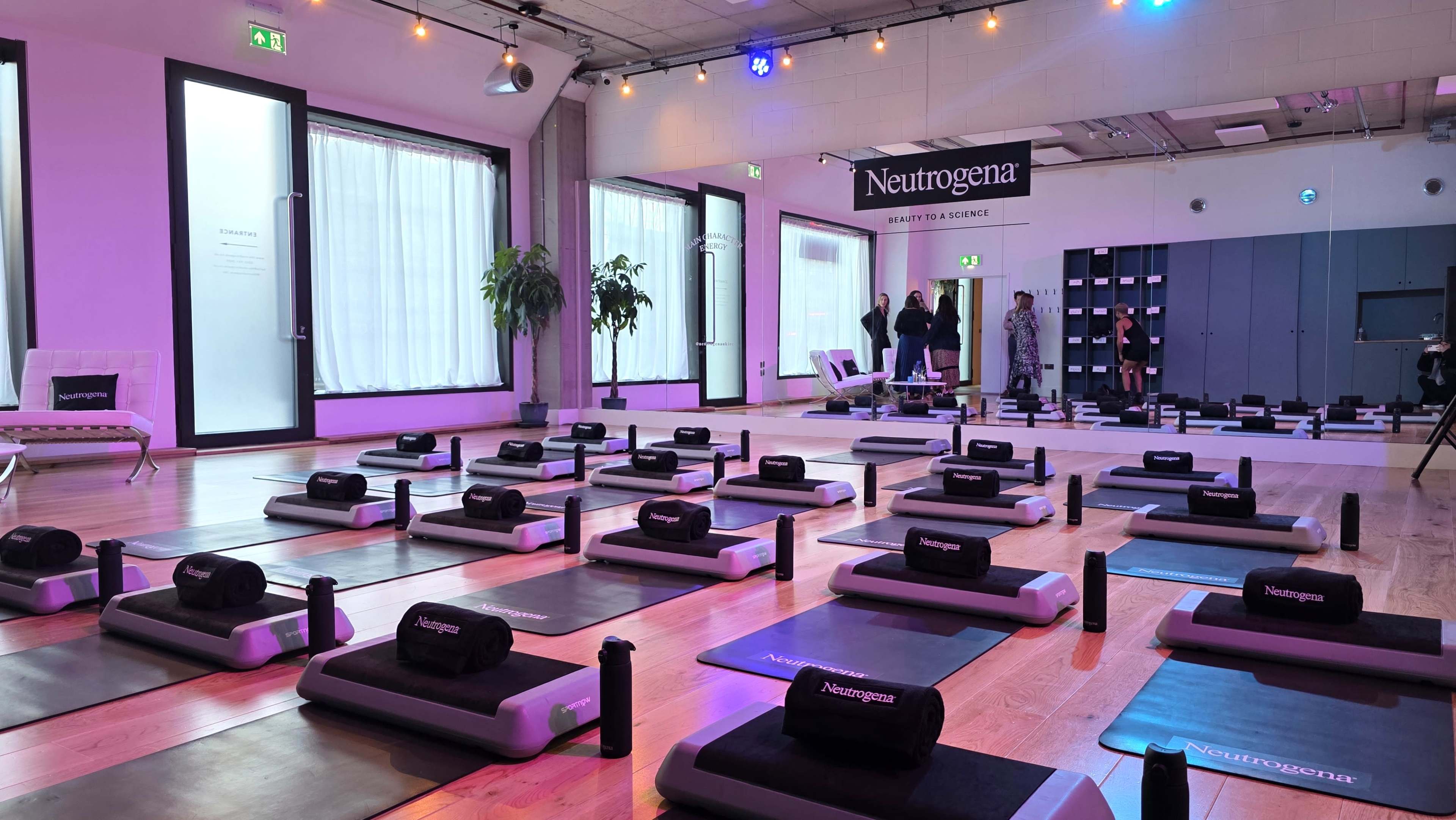 The image shows a brightly lit yoga studio with neatly arranged exercise mats and water bottles, and a banner for Neutrogena prominently displayed in the background.
