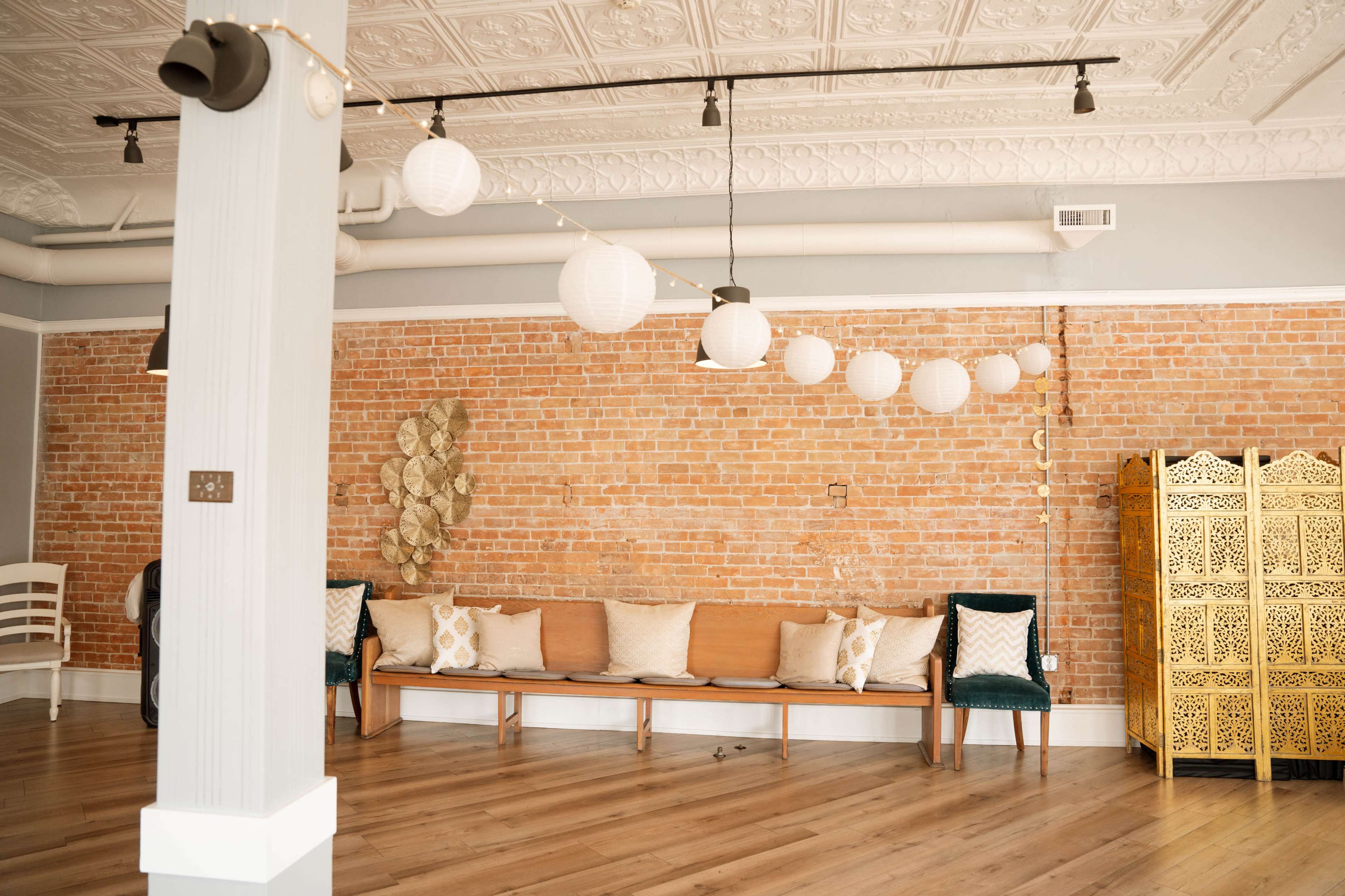 The image shows a spacious interior with a brick wall, wooden flooring, and a seating area featuring benches and cushions under decorative hanging lights.