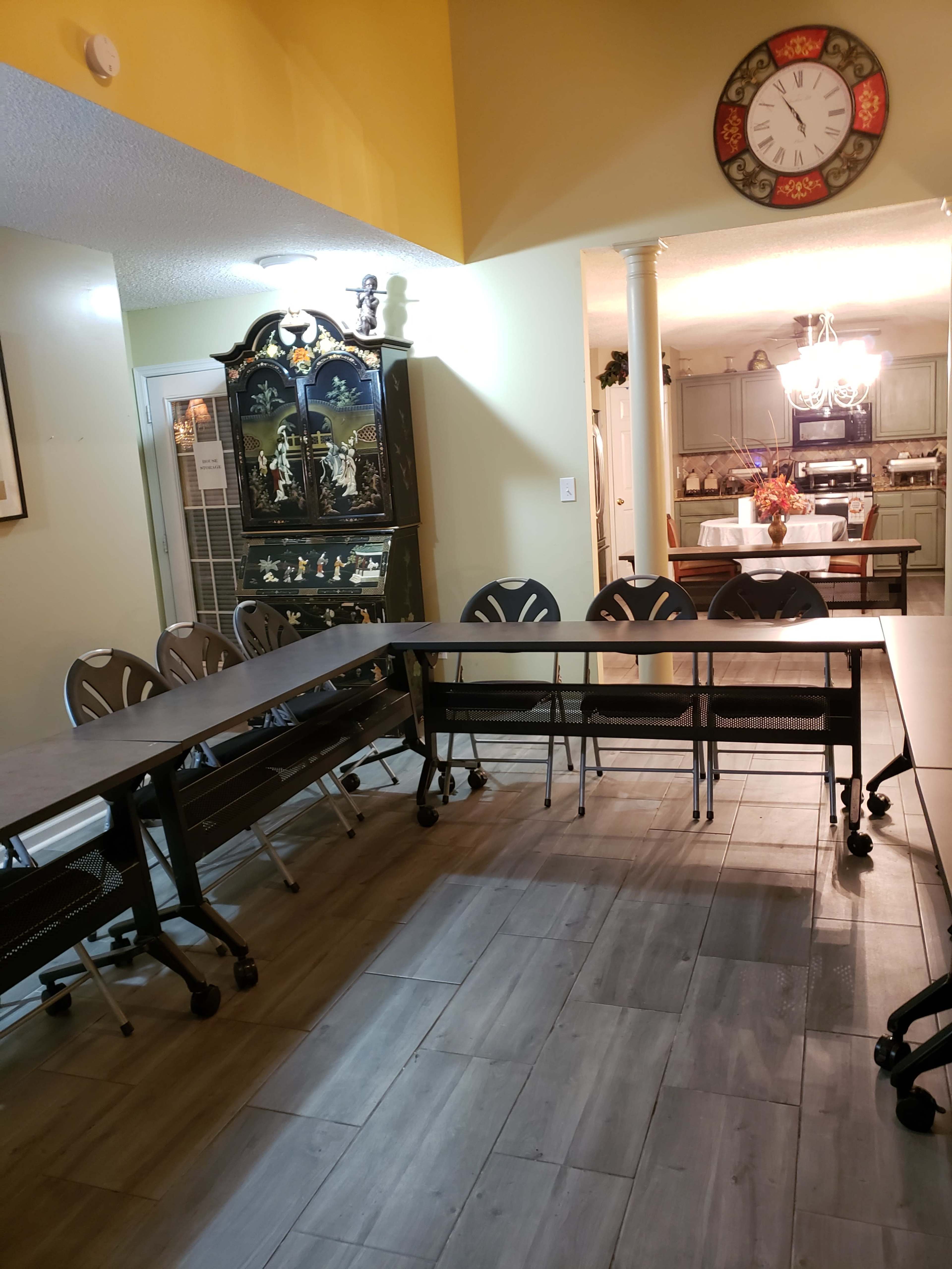 A large, rectangular table is set up with several rolling chairs around it in a well-lit room featuring a decorative cabinet and a kitchen area visible in the background.