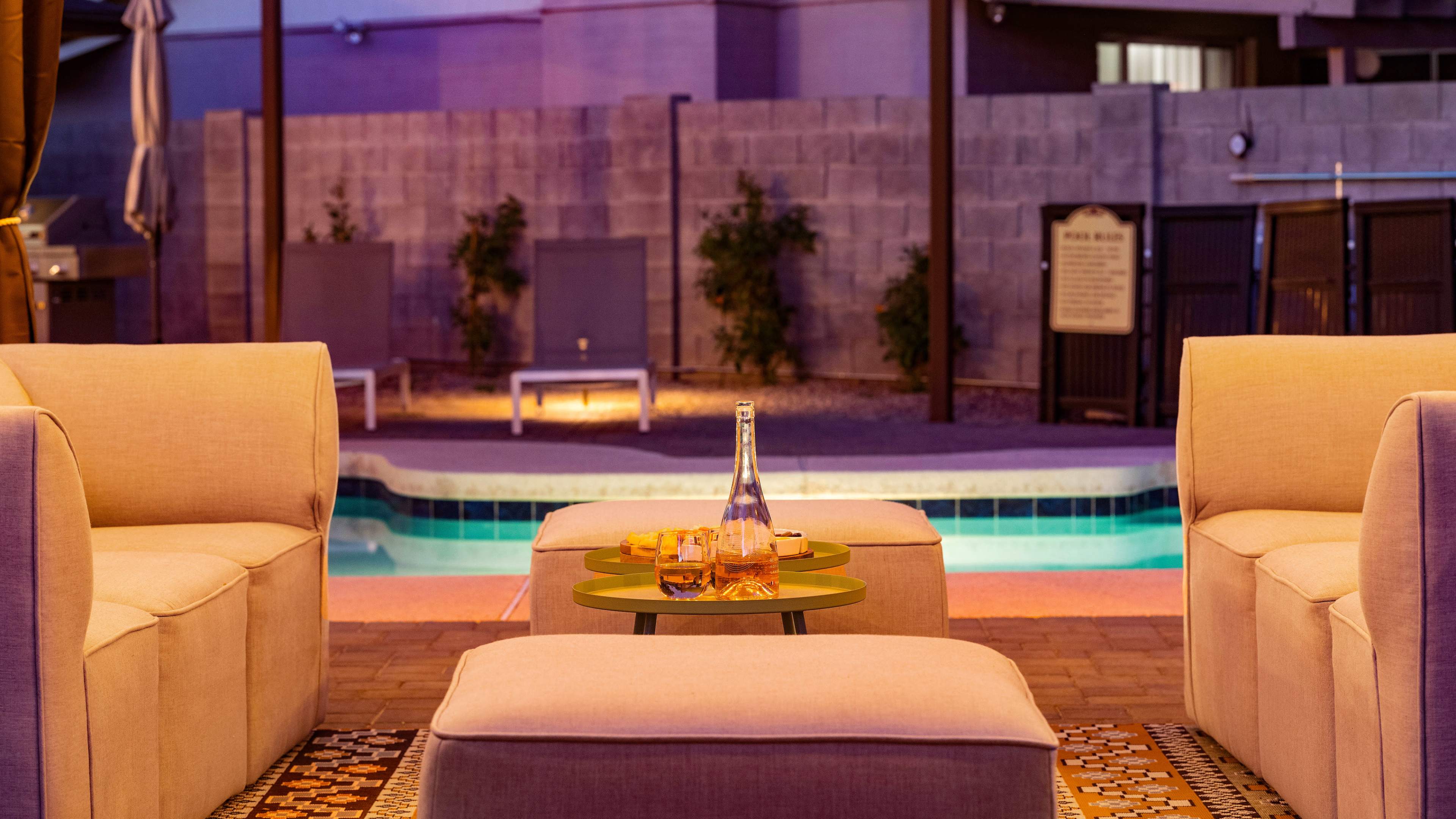 A small outdoor seating area with four light-colored chairs surrounding a green table, featuring a bottle and glasses, next to a pool illuminated by soft lighting.
