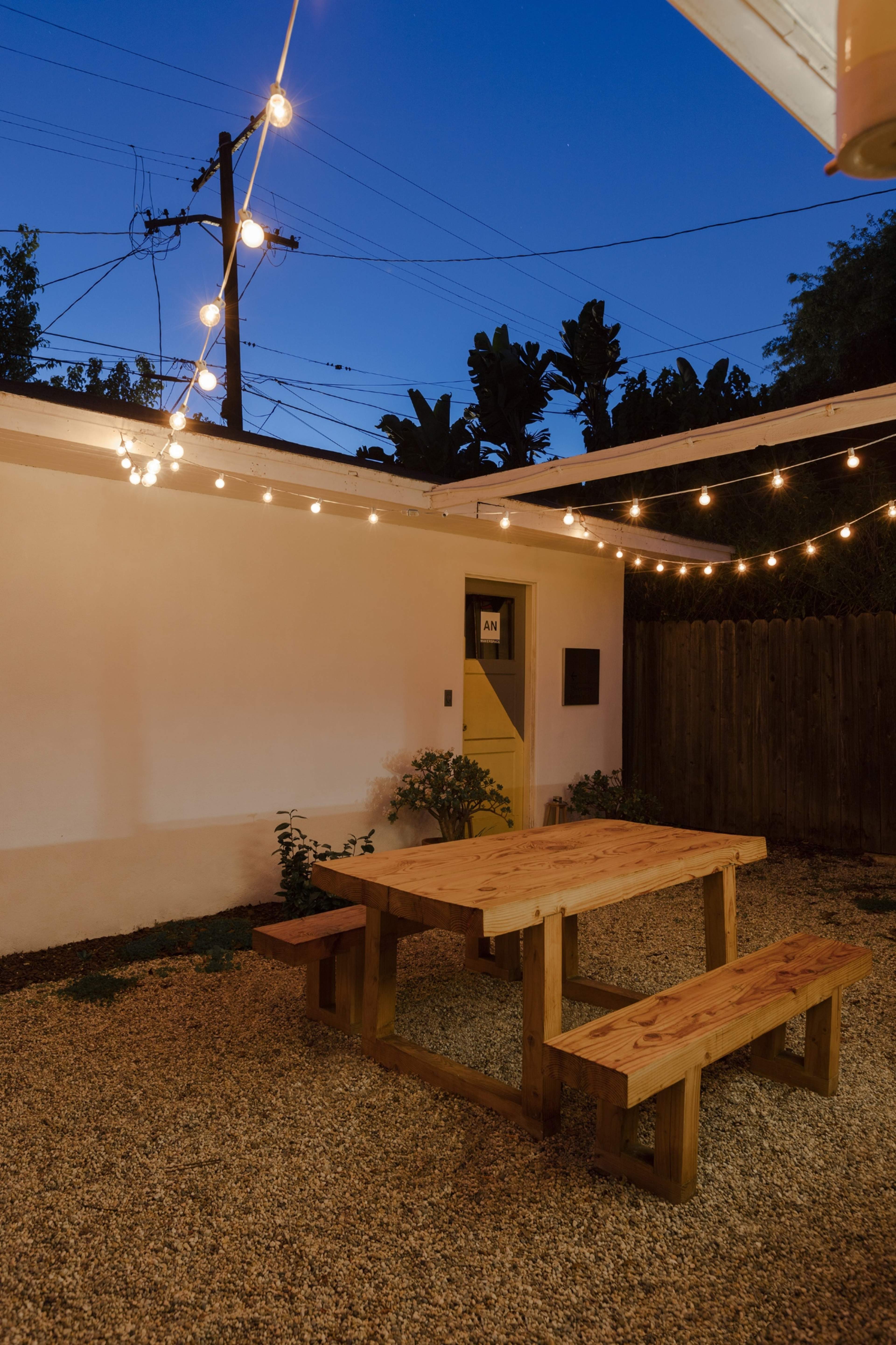 A wooden table and benches are set on a gravel surface beneath string lights, with a white wall and a door in the background.