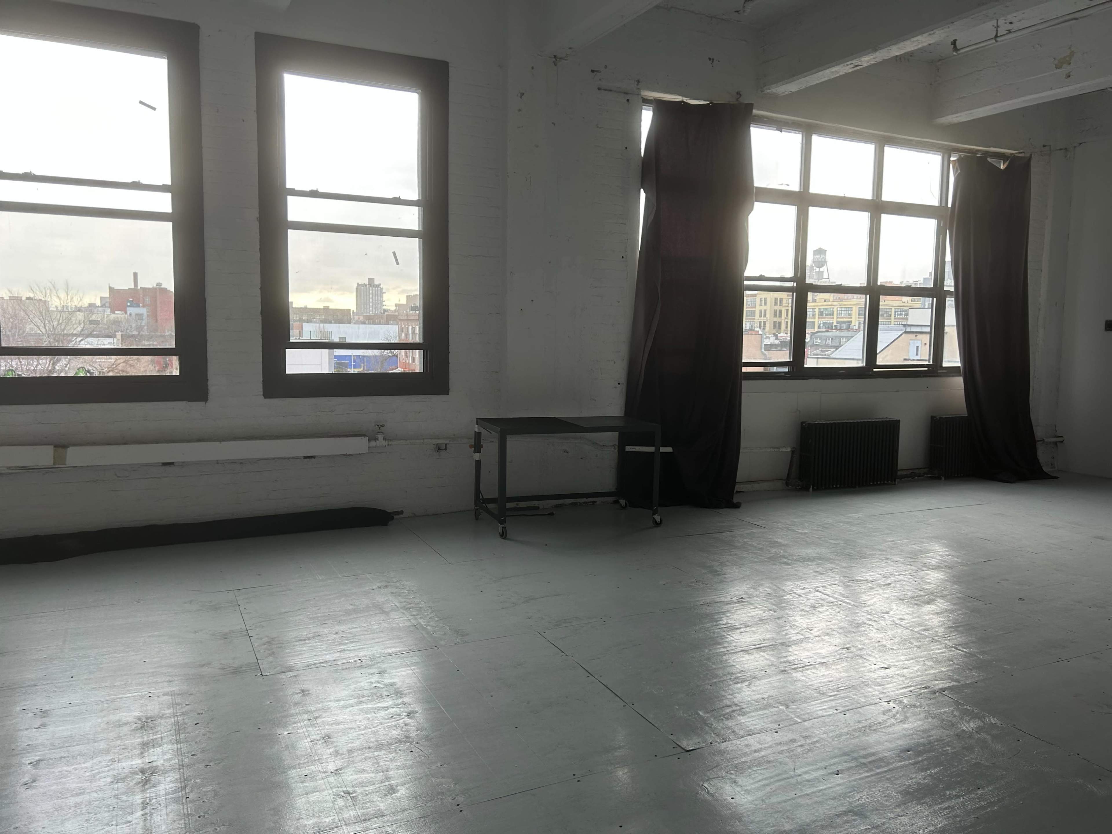 The image shows an empty room with white walls, large windows, and a table near the window.