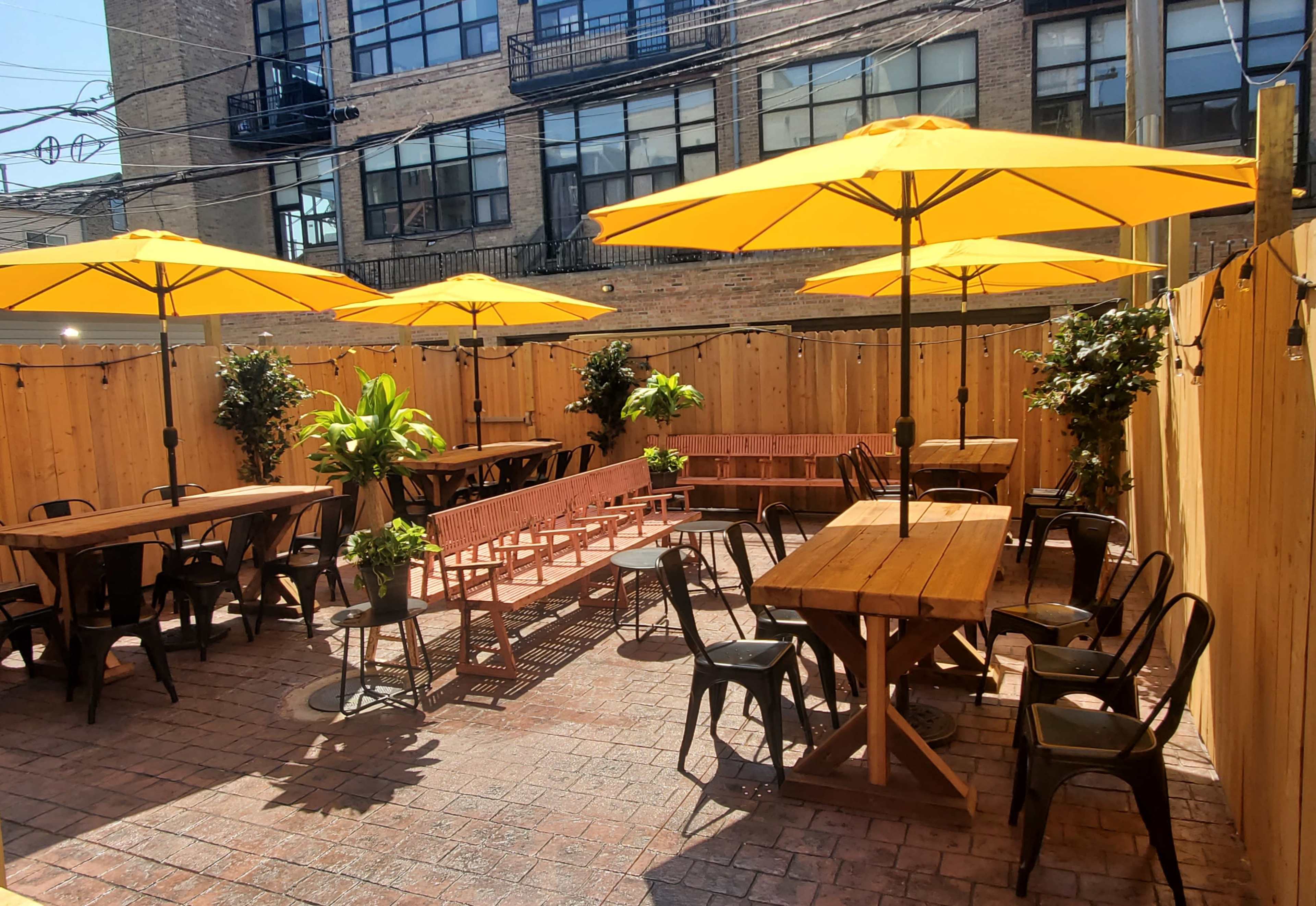 A courtyard with several wooden tables and yellow umbrellas surrounded by wooden fencing and greenery.