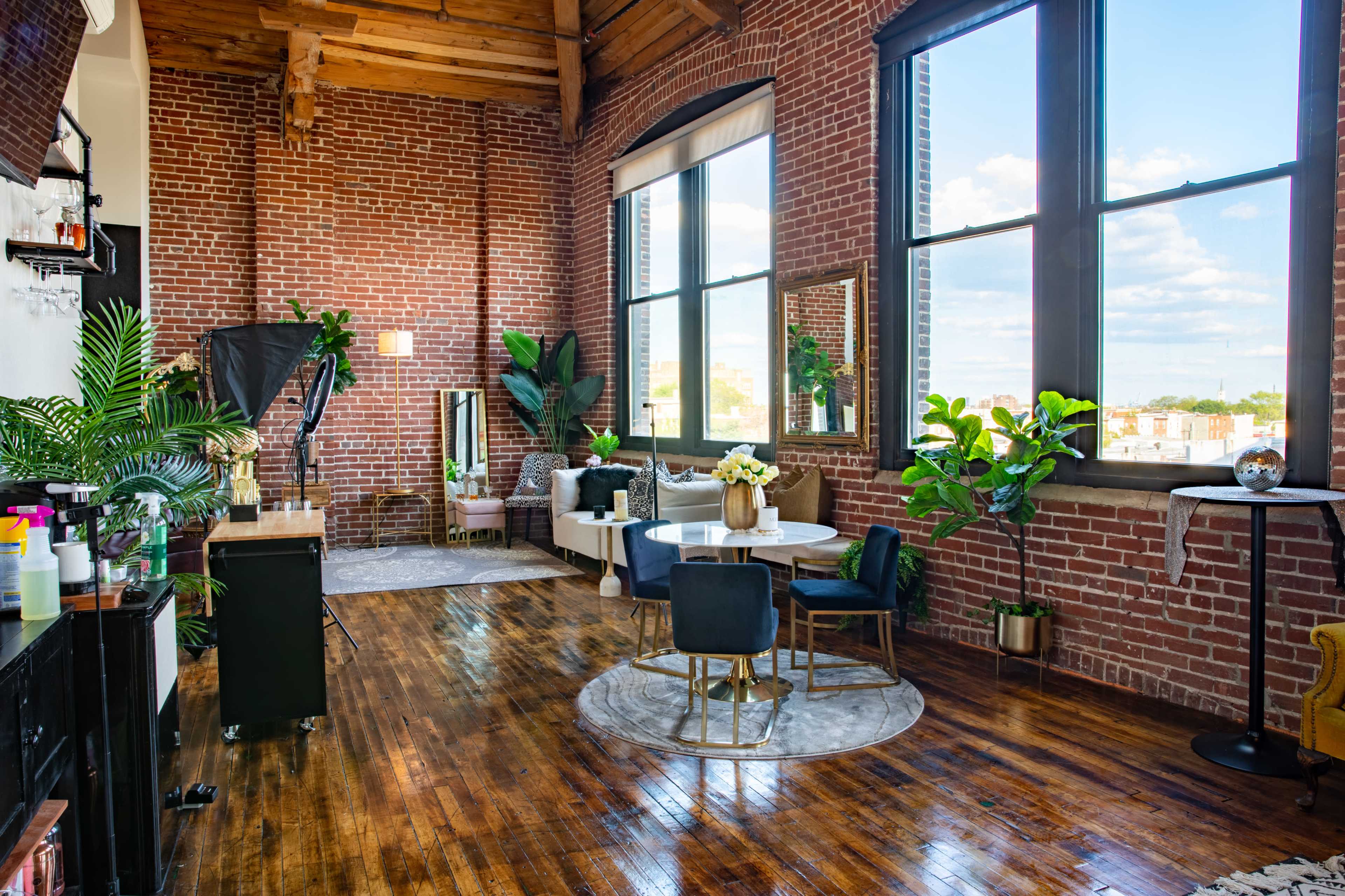 A spacious, industrial-style loft with exposed brick walls, large windows, and wooden floors, furnished with a seating area and plants.