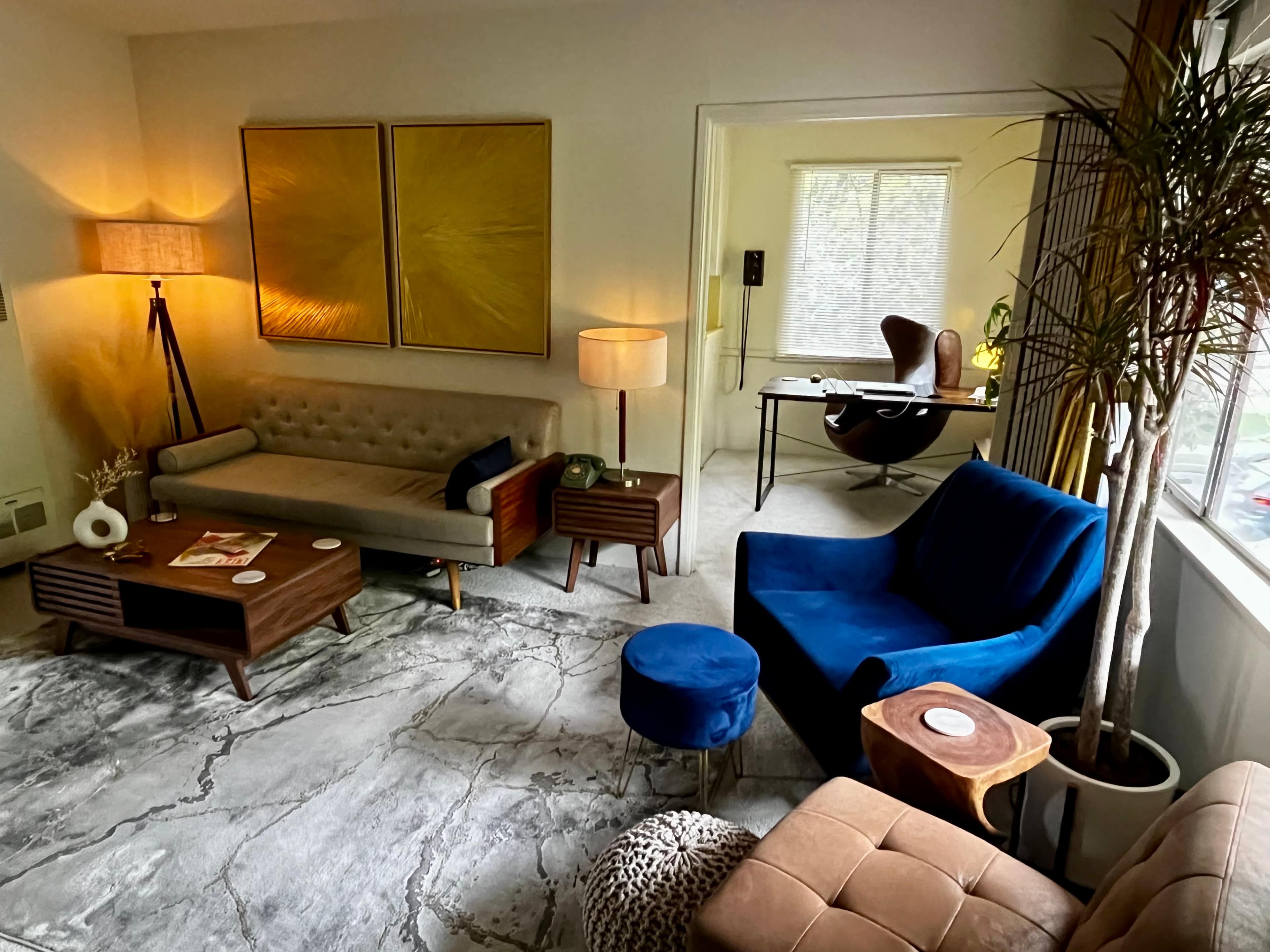A stylish living space featuring a beige sofa, a blue velvet chair, a wooden coffee table, and a desk in an adjacent room.
