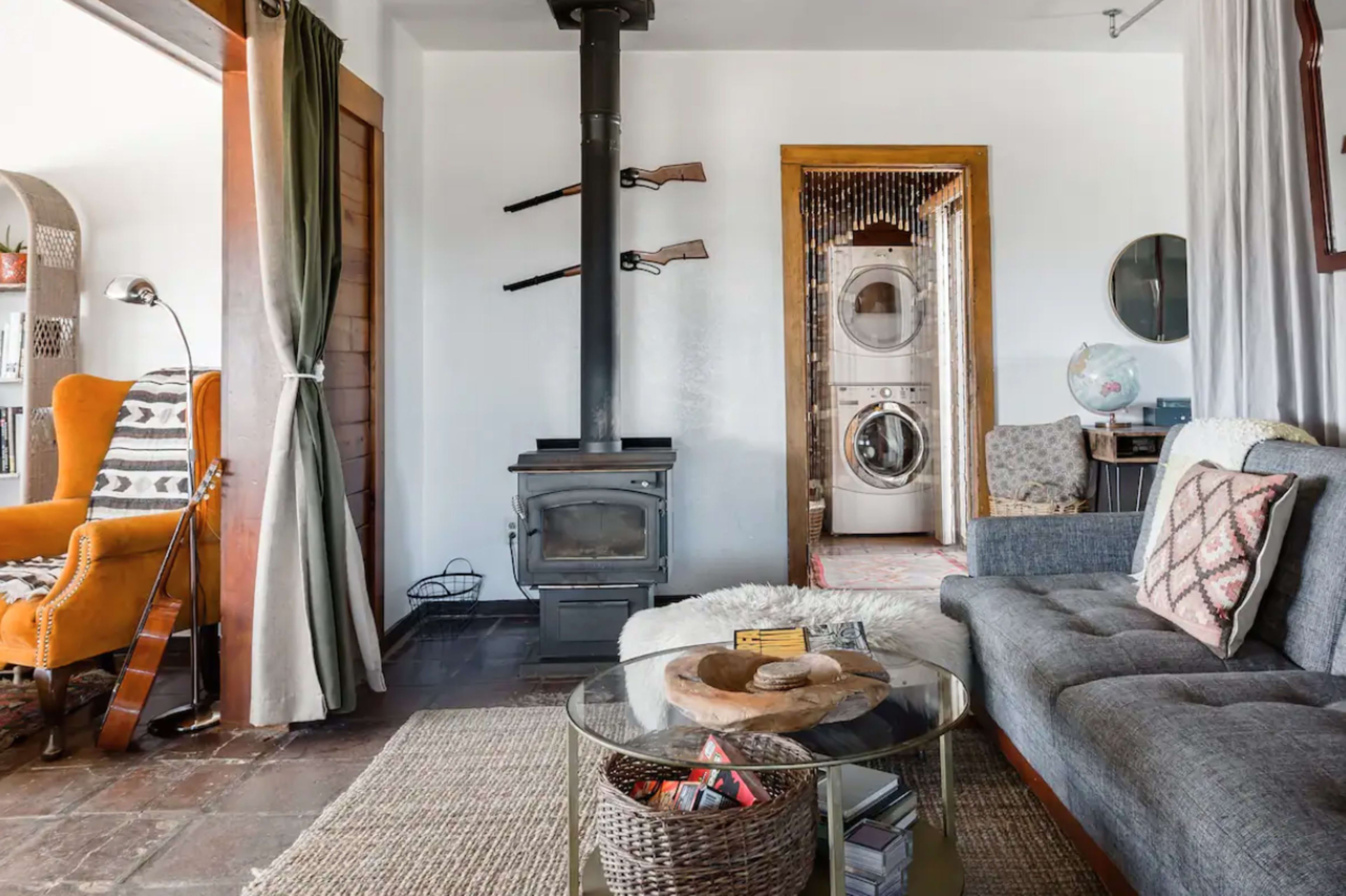 A living room with a wood stove, a gray sofa, and an orange armchair, along with a washing machine visible in the background.