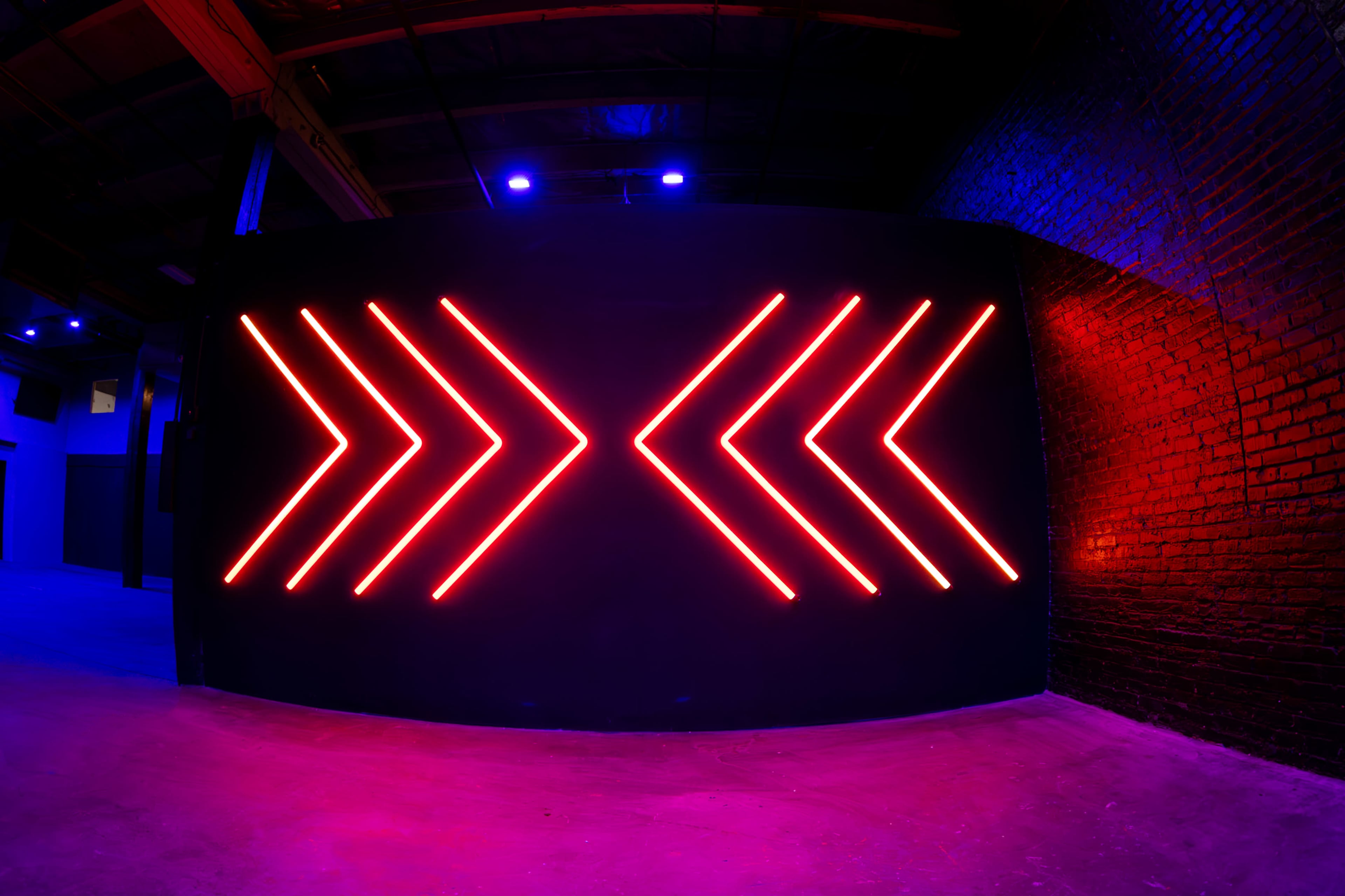The image shows a dark interior space featuring a wall with vibrant red neon arrow shapes arranged in a symmetrical pattern.