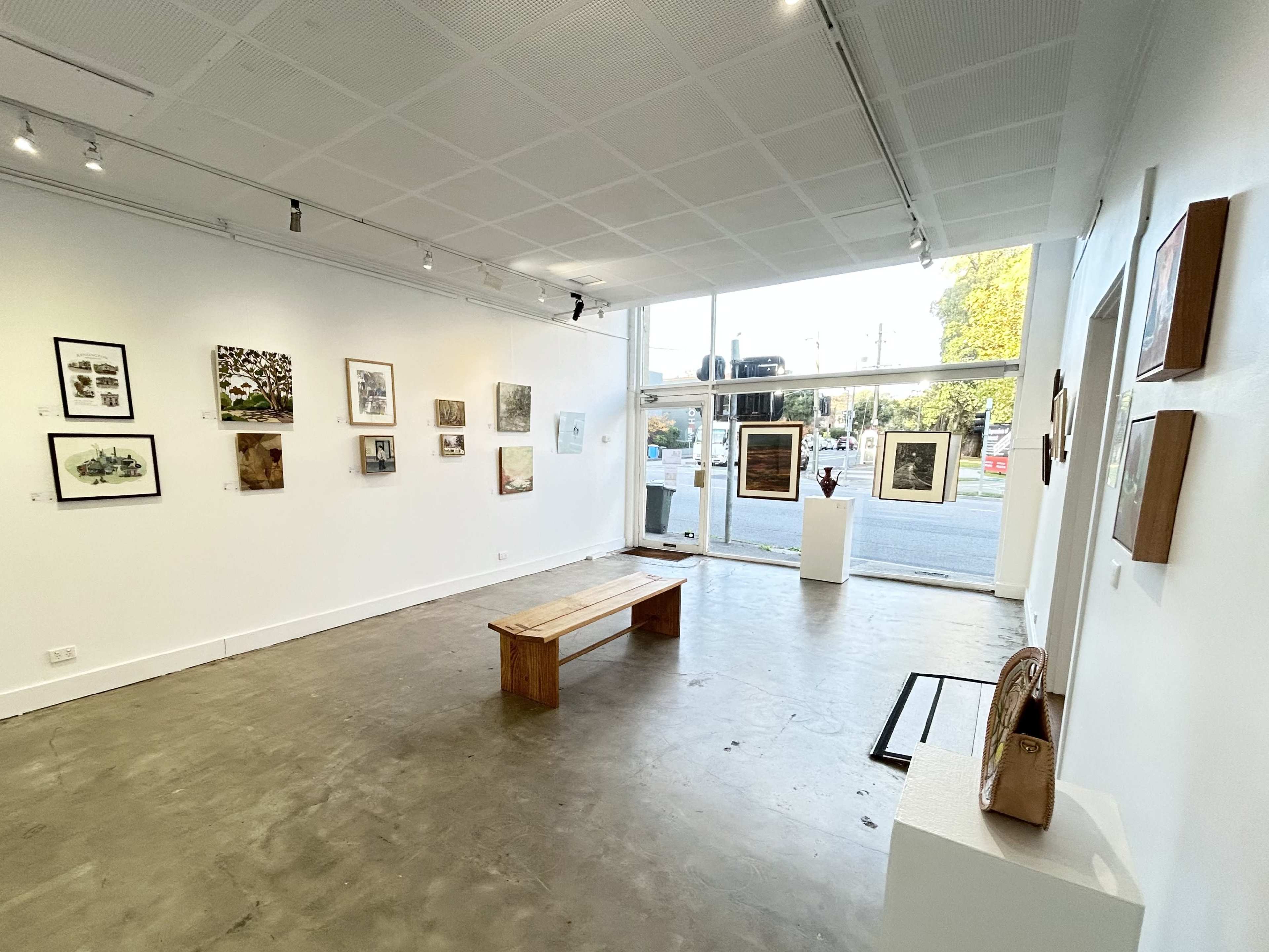 The image shows a contemporary art gallery with white walls displaying various framed artworks and a wooden bench in the center.