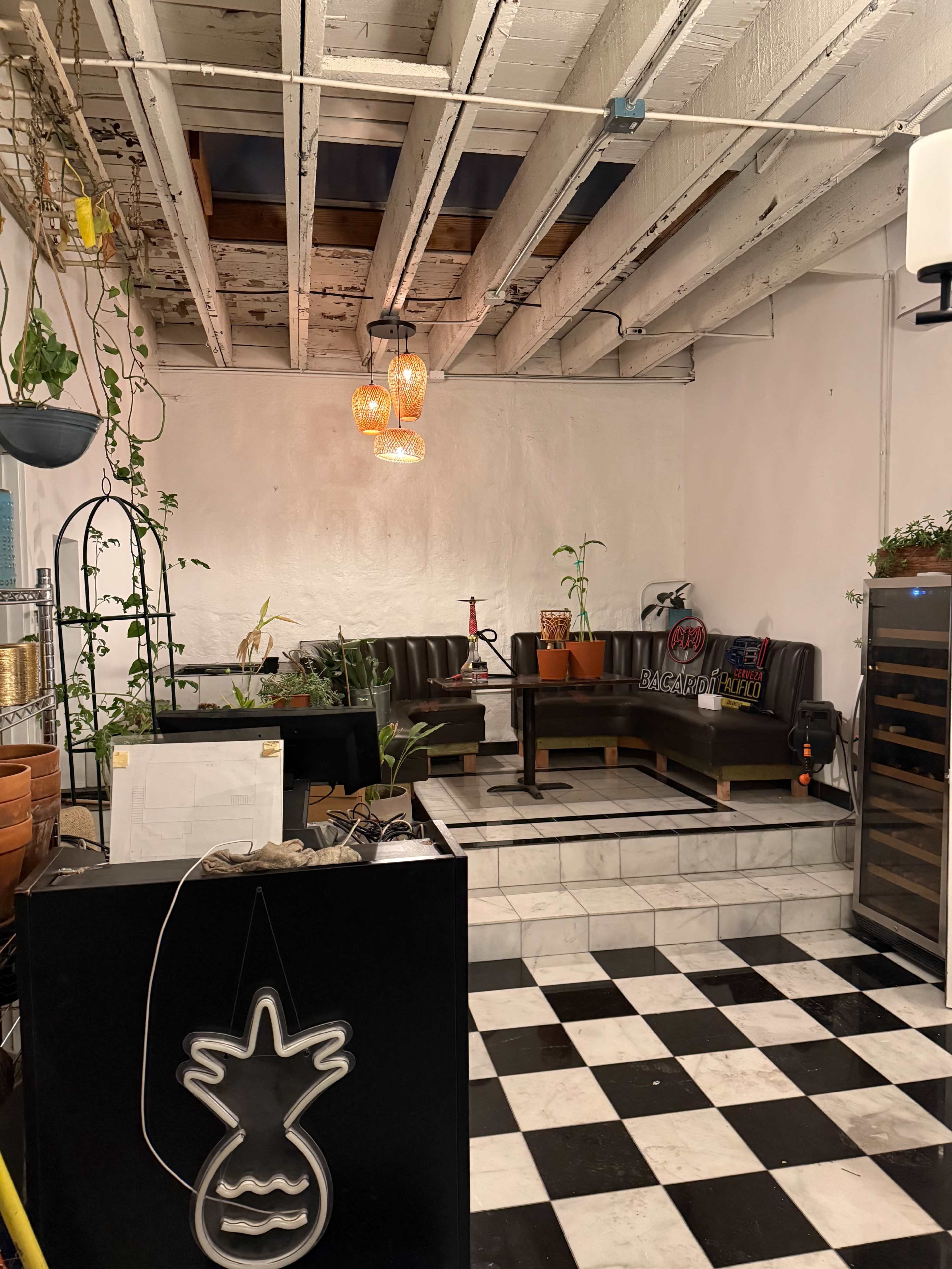 The image shows a cozy cafe interior featuring black and white checkered flooring, a sectional sofa, a coffee table surrounded by plants, and decorative pendant lights.