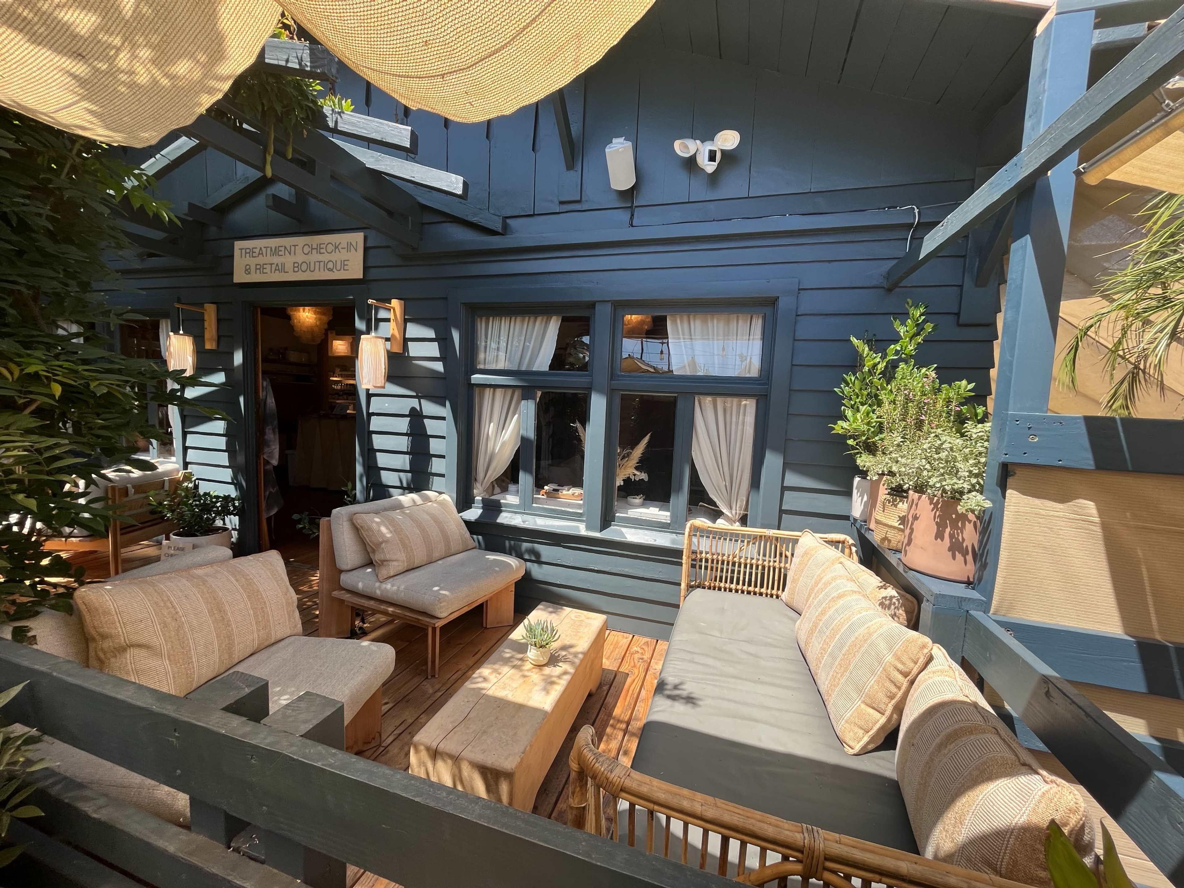 A cozy outdoor seating area with a wooden table and comfortable couches, adjacent to a blue building with large windows and greenery.