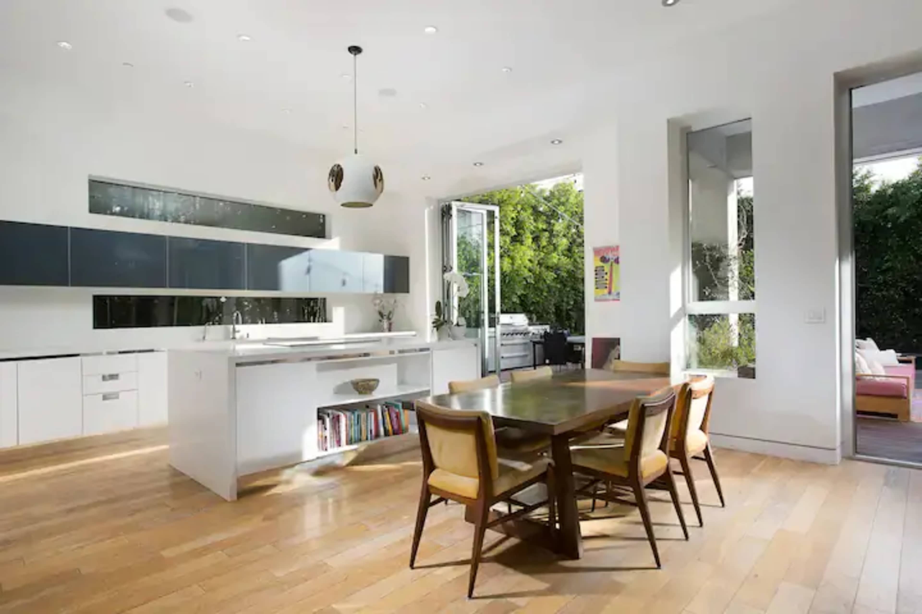 A modern kitchen features white cabinetry, a central island, and a dining table surrounded by four chairs, with large windows opening to a garden.