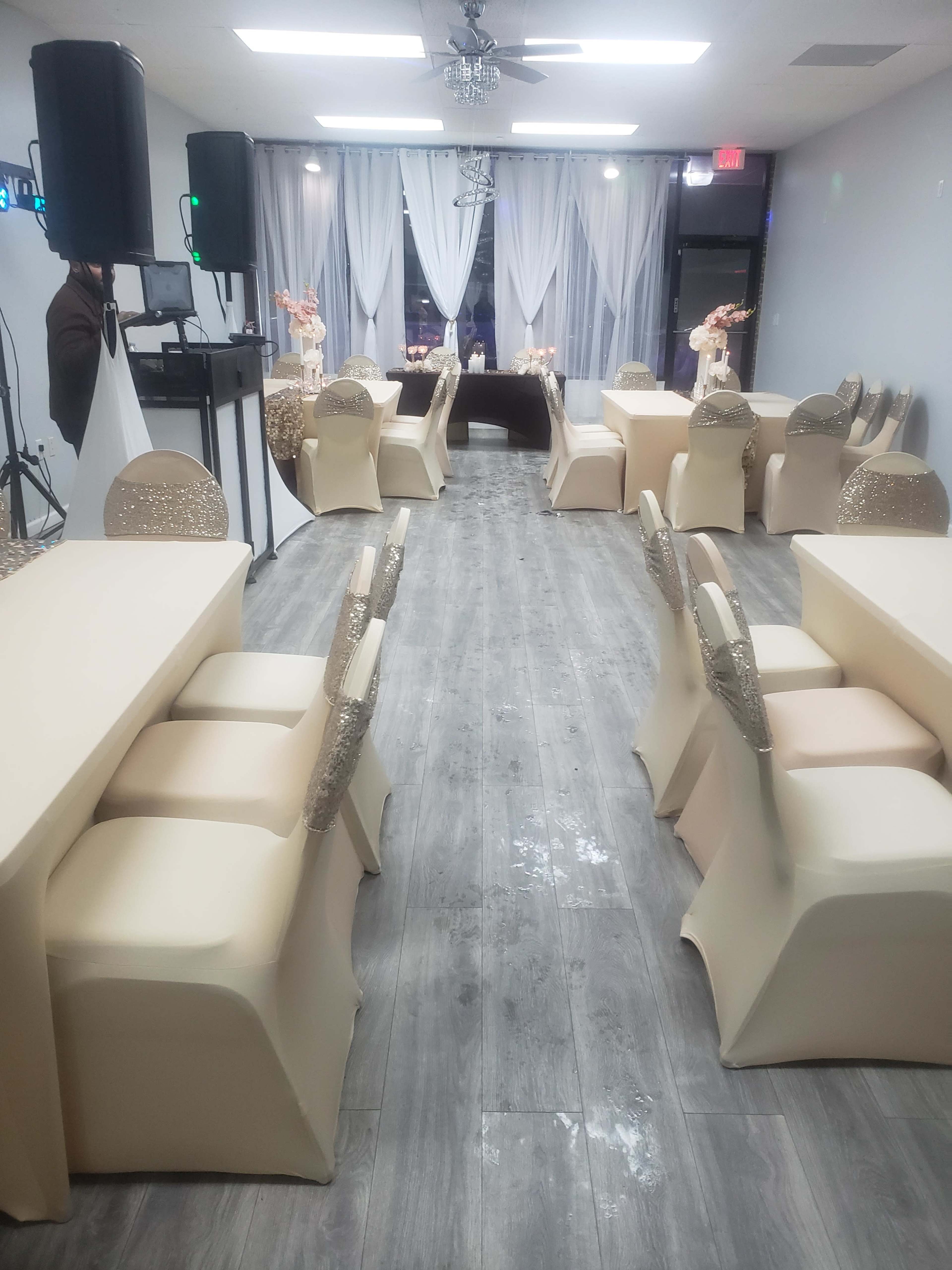 A spacious event room features neatly arranged tables with beige tablecloths and decorated chairs, illuminated by soft lighting and adorned with floral centerpieces.