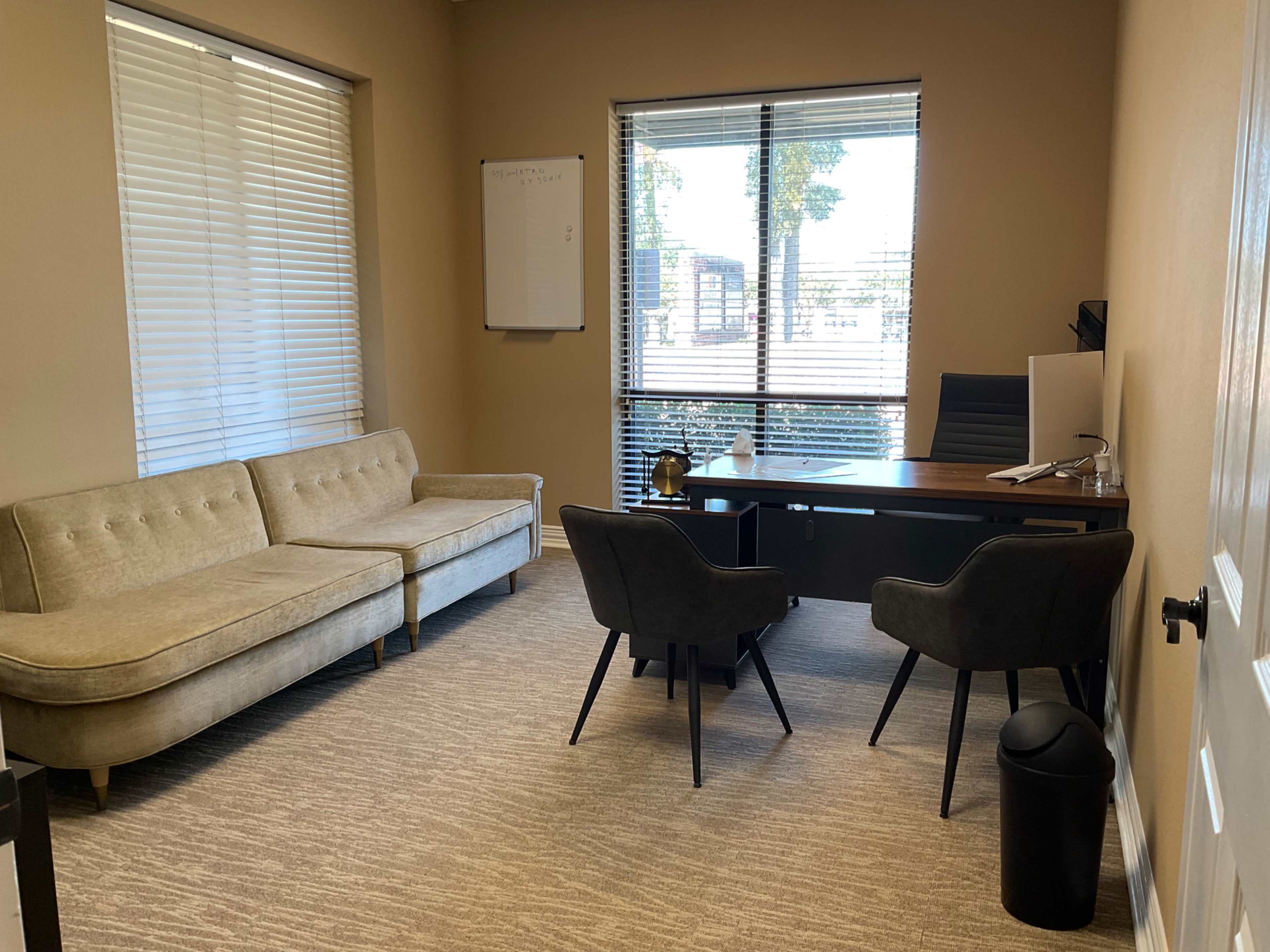 The image shows a small office with a beige sofa, two black chairs, a wooden desk with a computer, and a window with blinds letting in natural light.