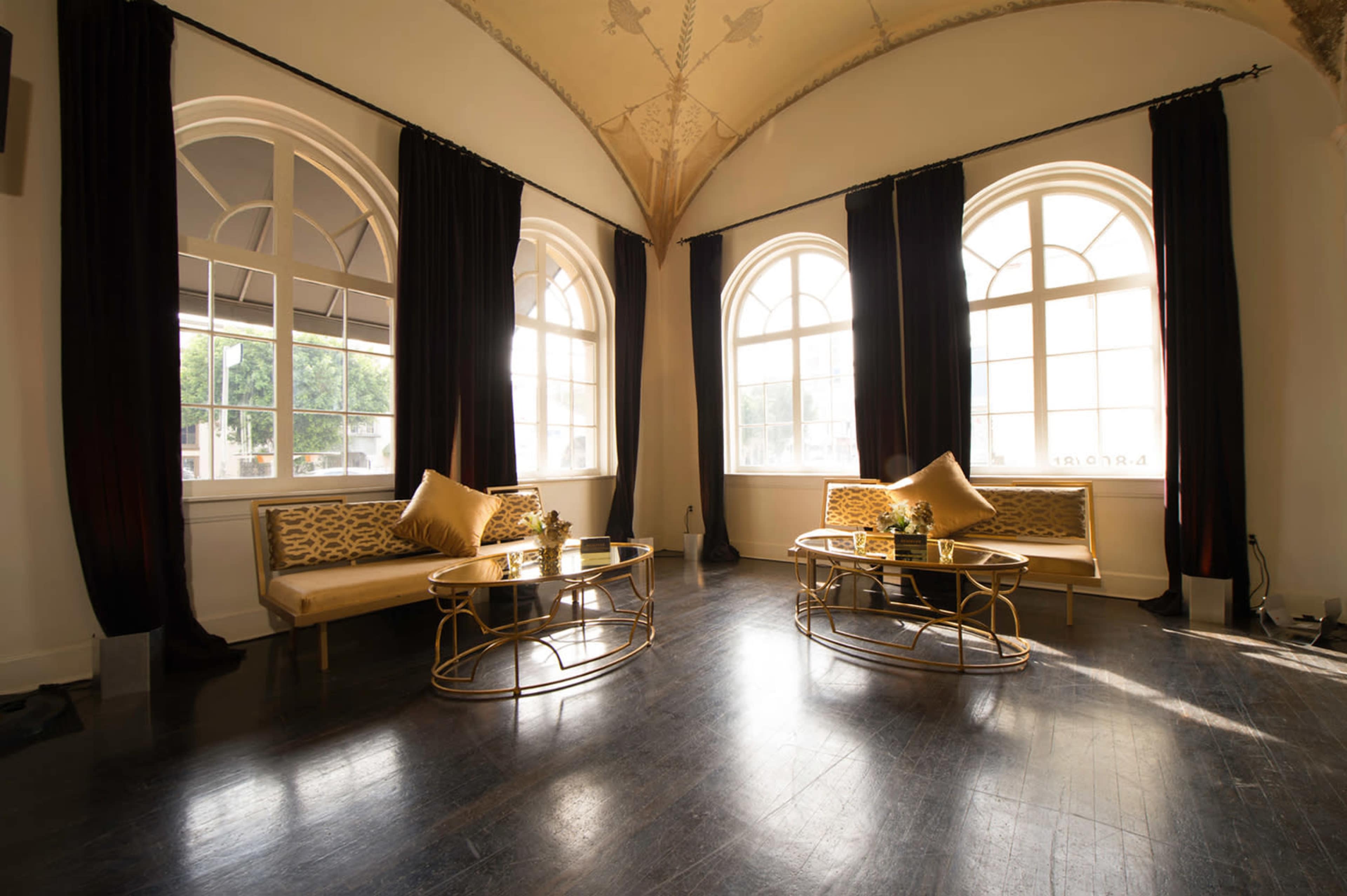 The image shows a stylish interior with two wooden sofas and gold tables set against large arched windows, illuminated by natural light.