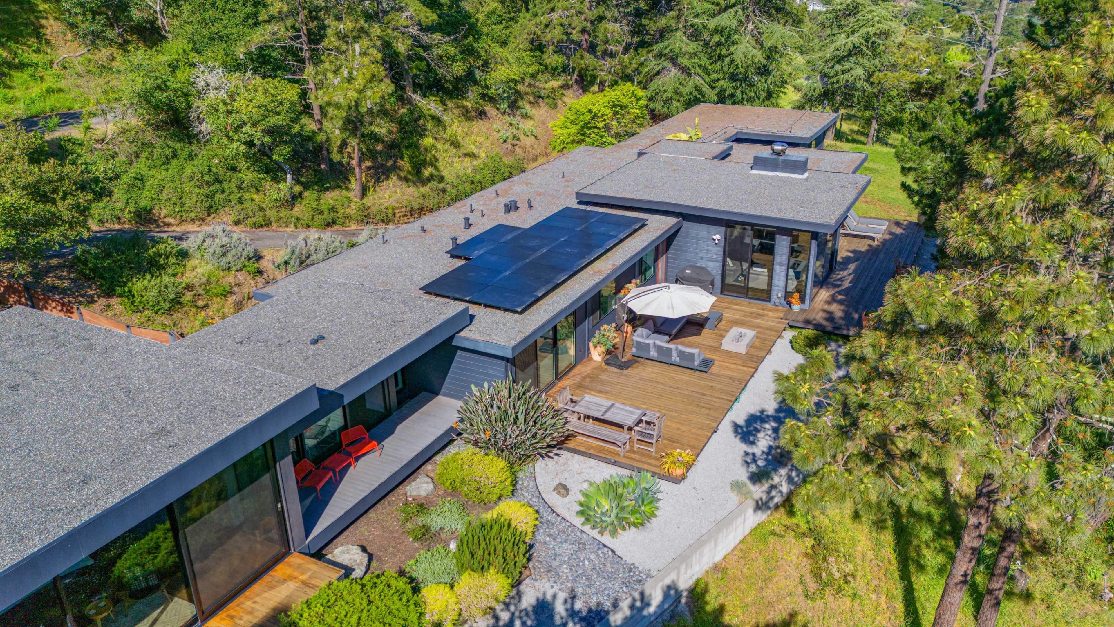 A modern single-story home features a flat roof with solar panels, surrounded by landscaped gardens and a wooden deck with outdoor seating.
