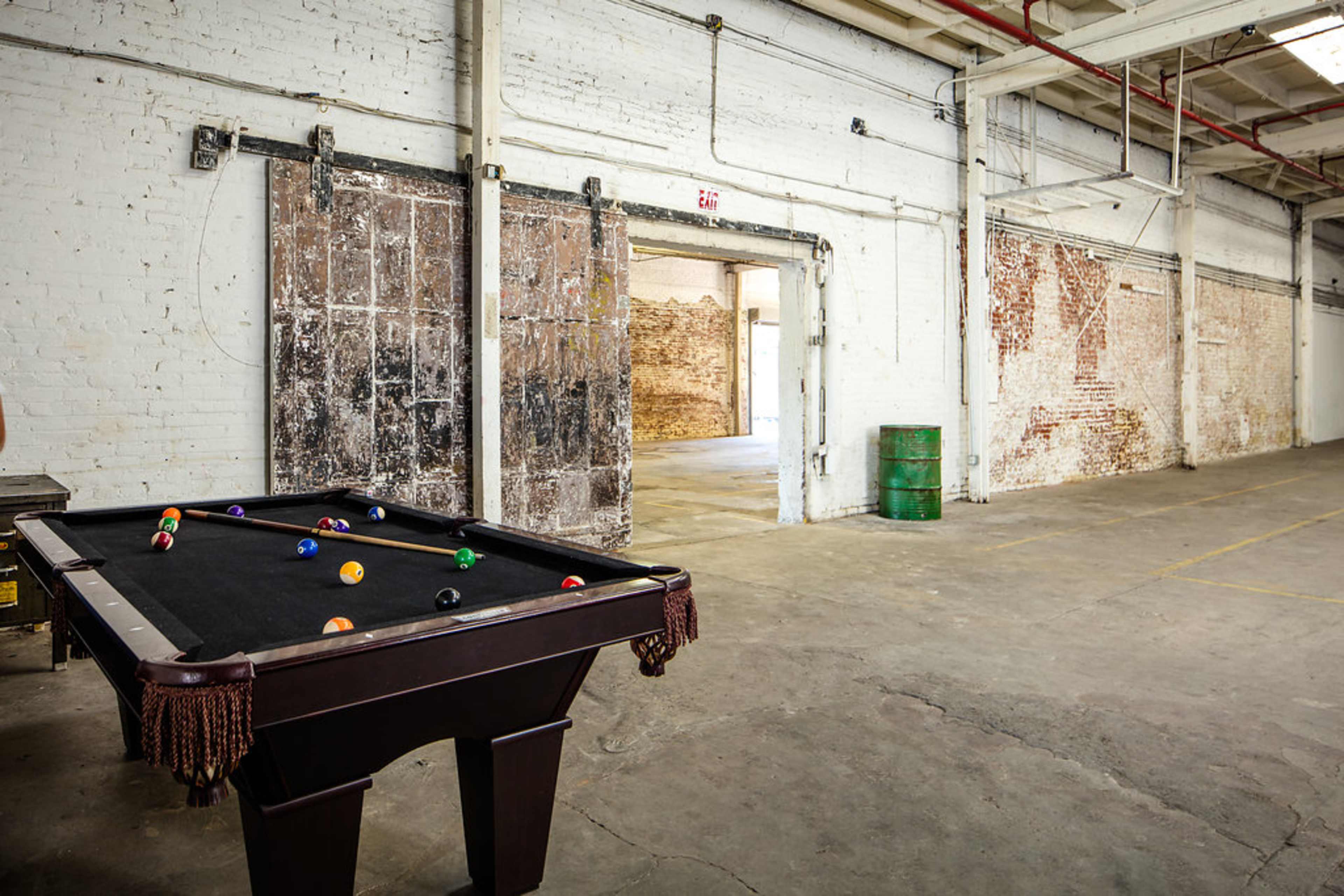 A pool table is positioned in an empty, industrial-style room with exposed brick walls and concrete flooring.
