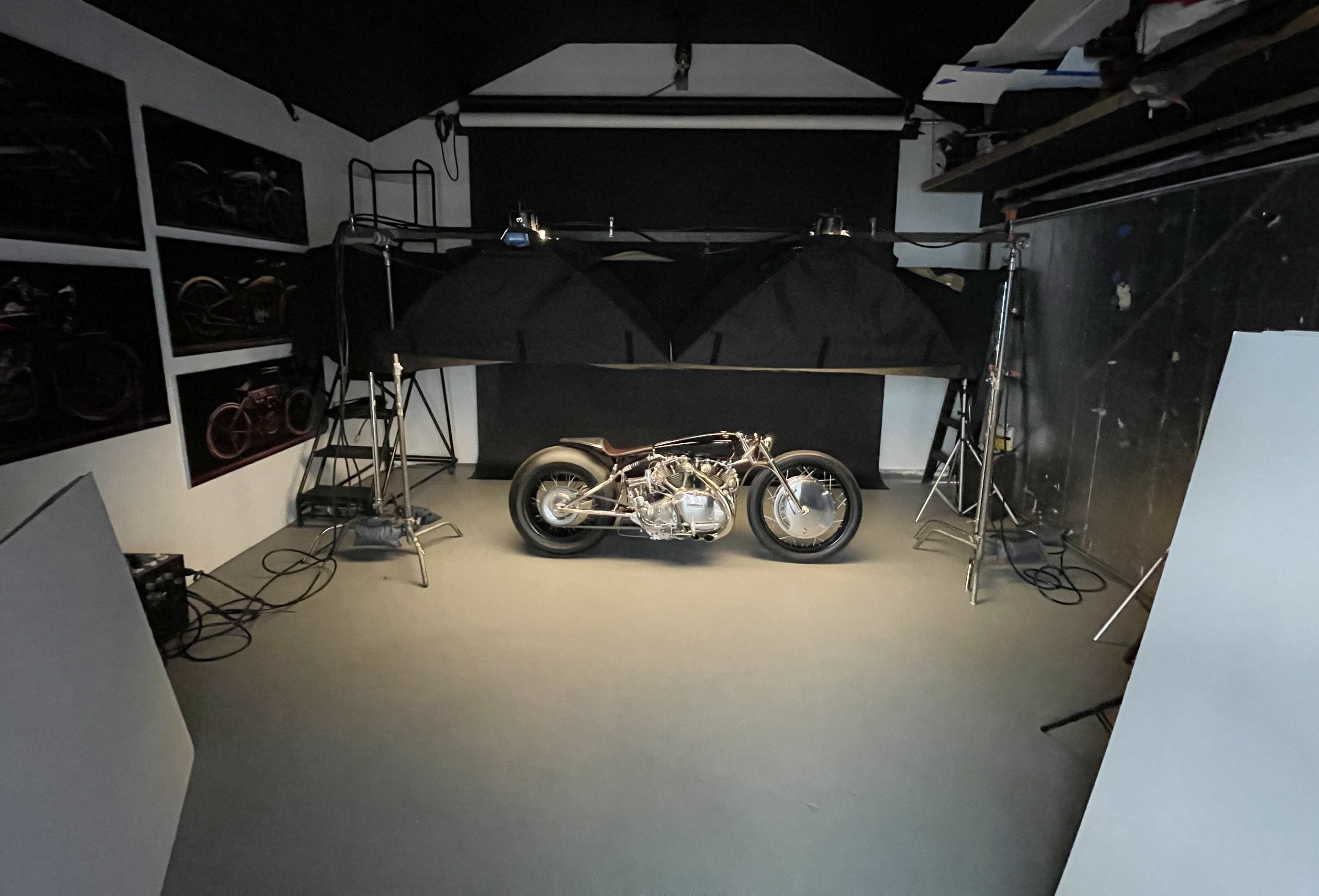 A motorcycle is positioned on a well-lit studio floor surrounded by lighting equipment and black backdrop drapes.