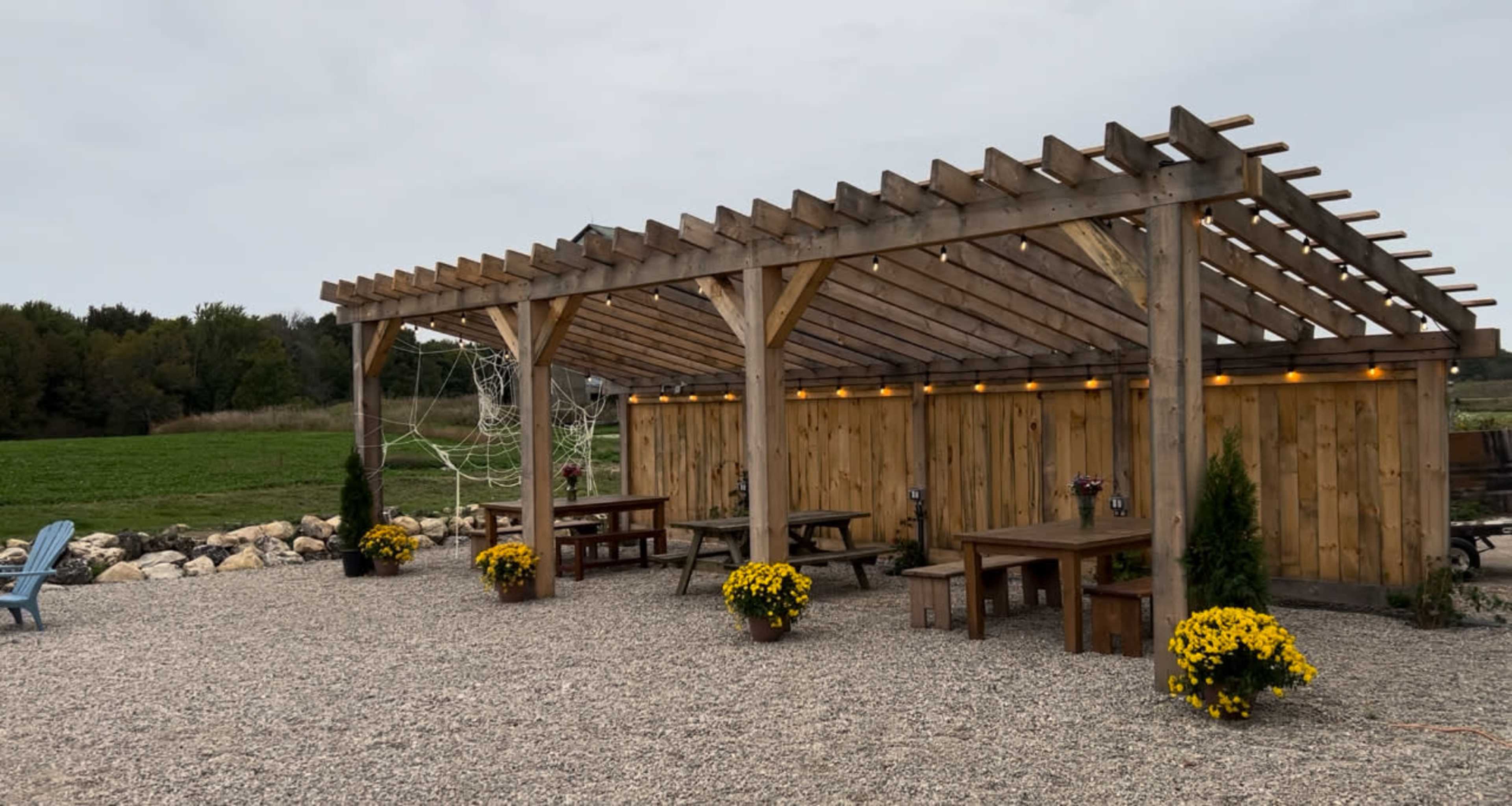 The image shows a wooden outdoor structure with a slatted roof, surrounded by tables and flower pots, situated near a grassy area and stone border.