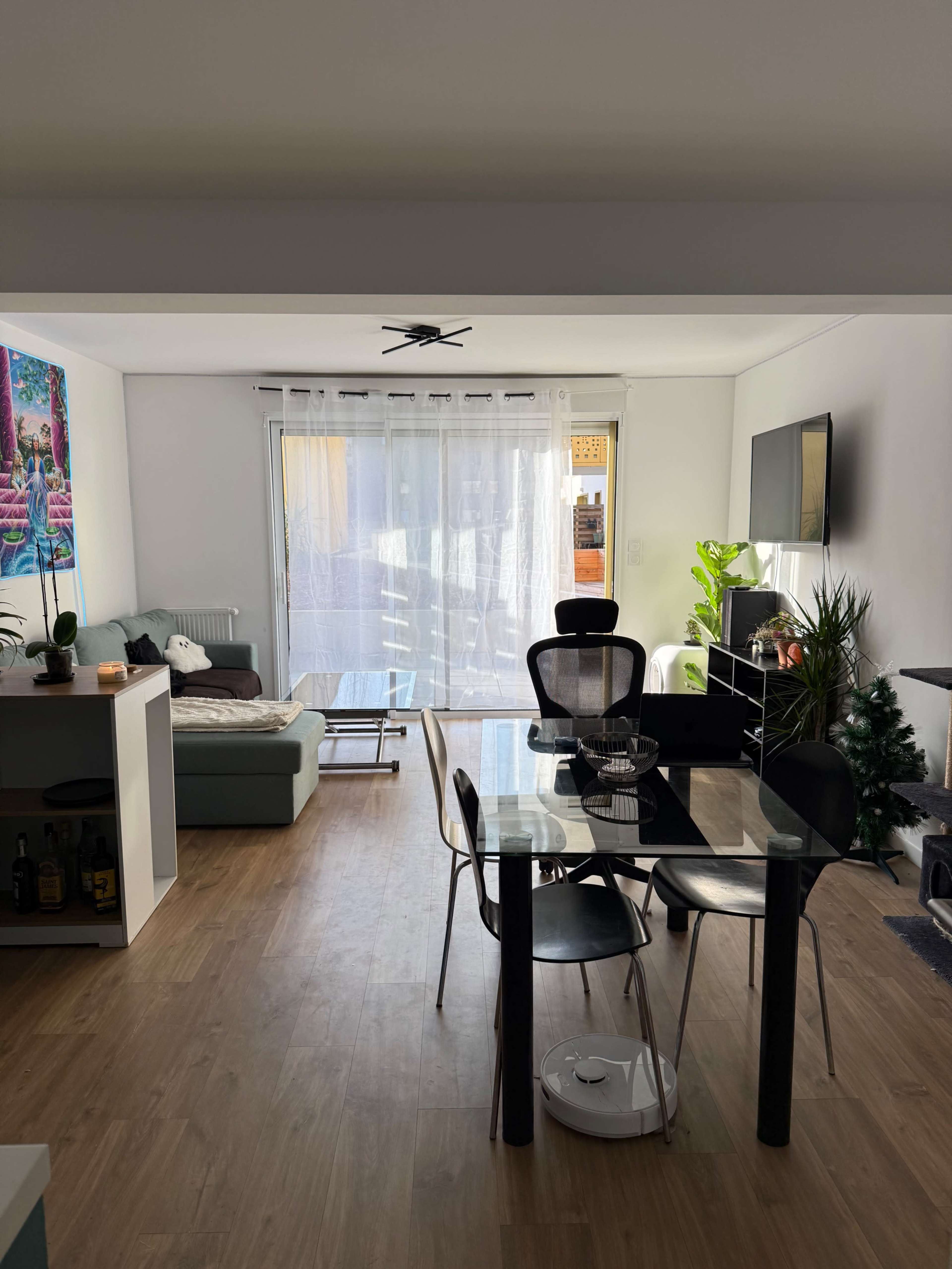 The image shows a bright living area with a dining table and chairs, a couch, a television, and a large window partially covered by sheer curtains.