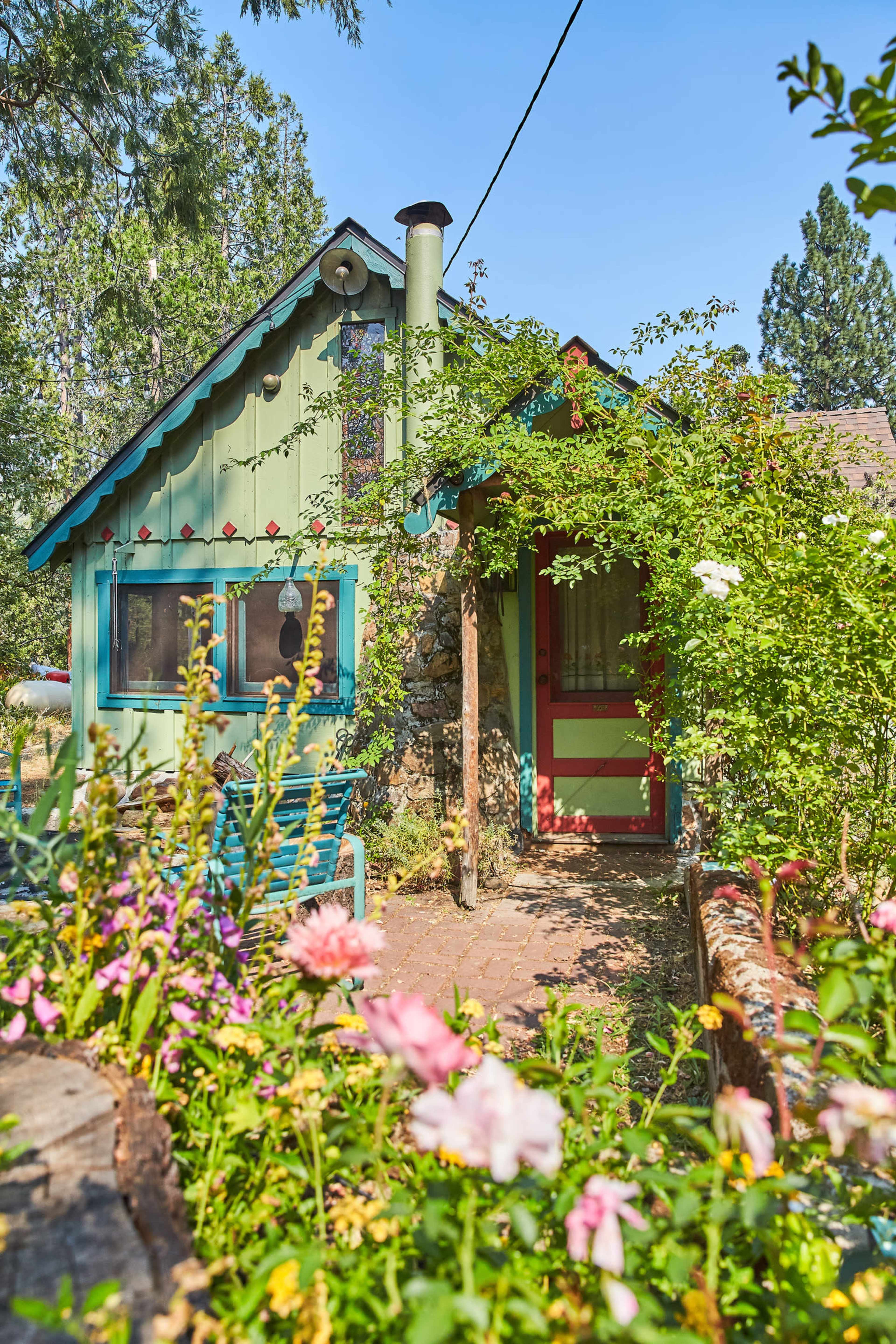 A small, brightly painted cottage is surrounded by colorful floral plants and greenery.