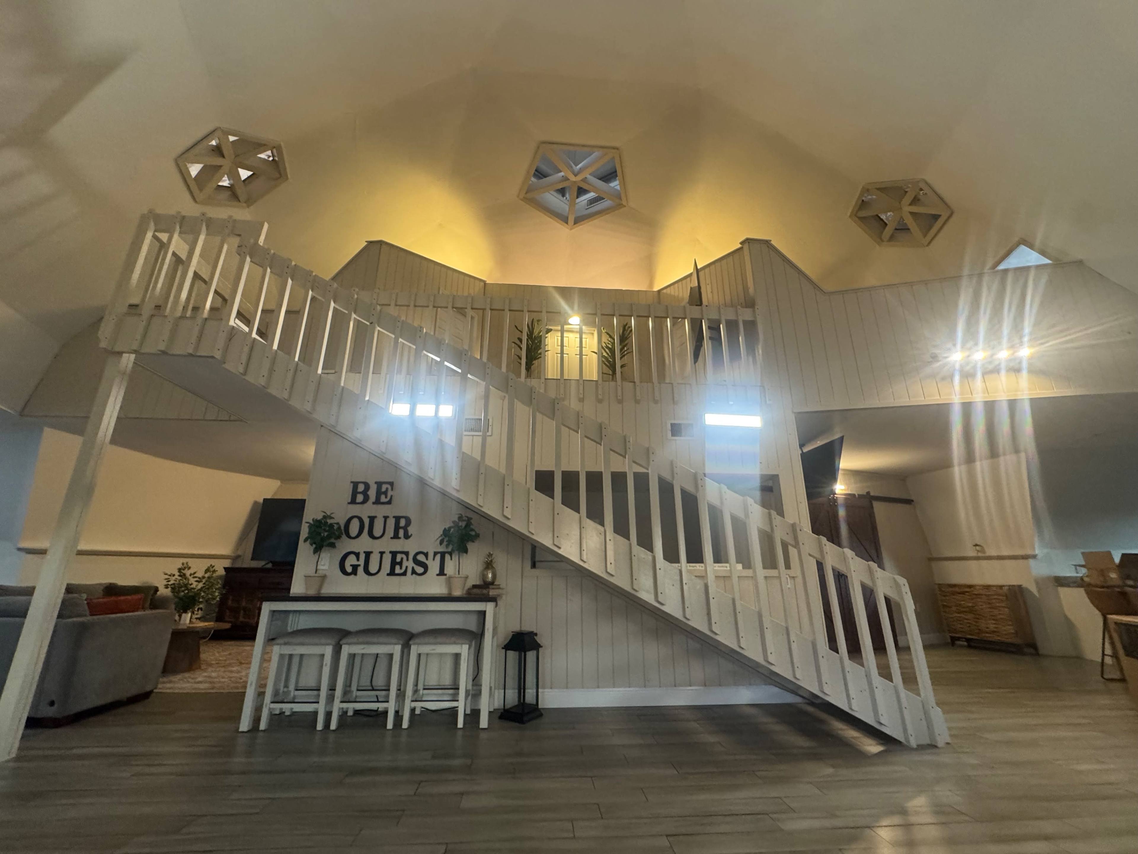 A spacious interior features a prominent staircase leading to a second level, with geometric decorations on the walls and a welcoming message displayed below.