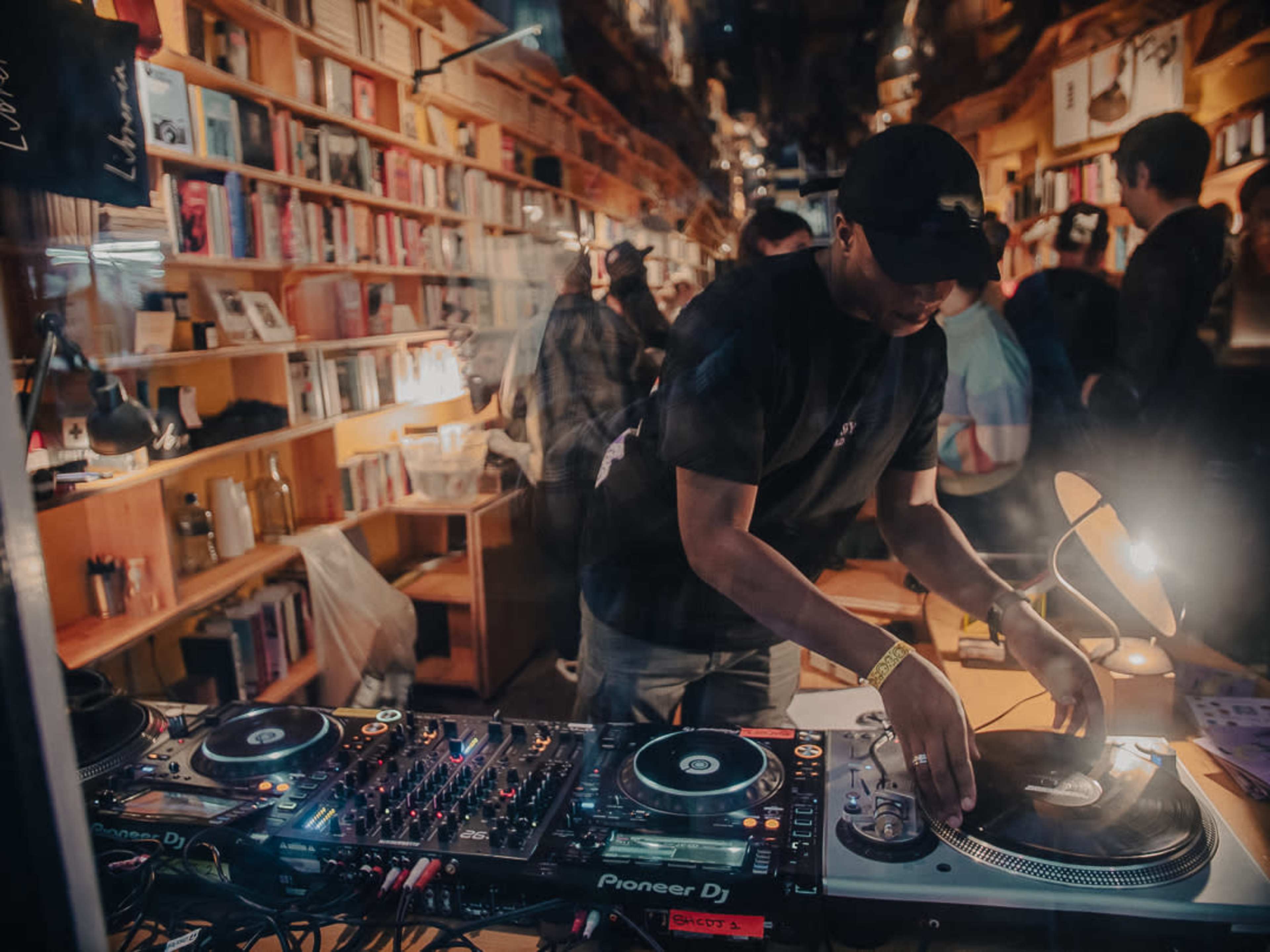 A DJ is mixing music at a turntable setup inside a crowded bookstore.
