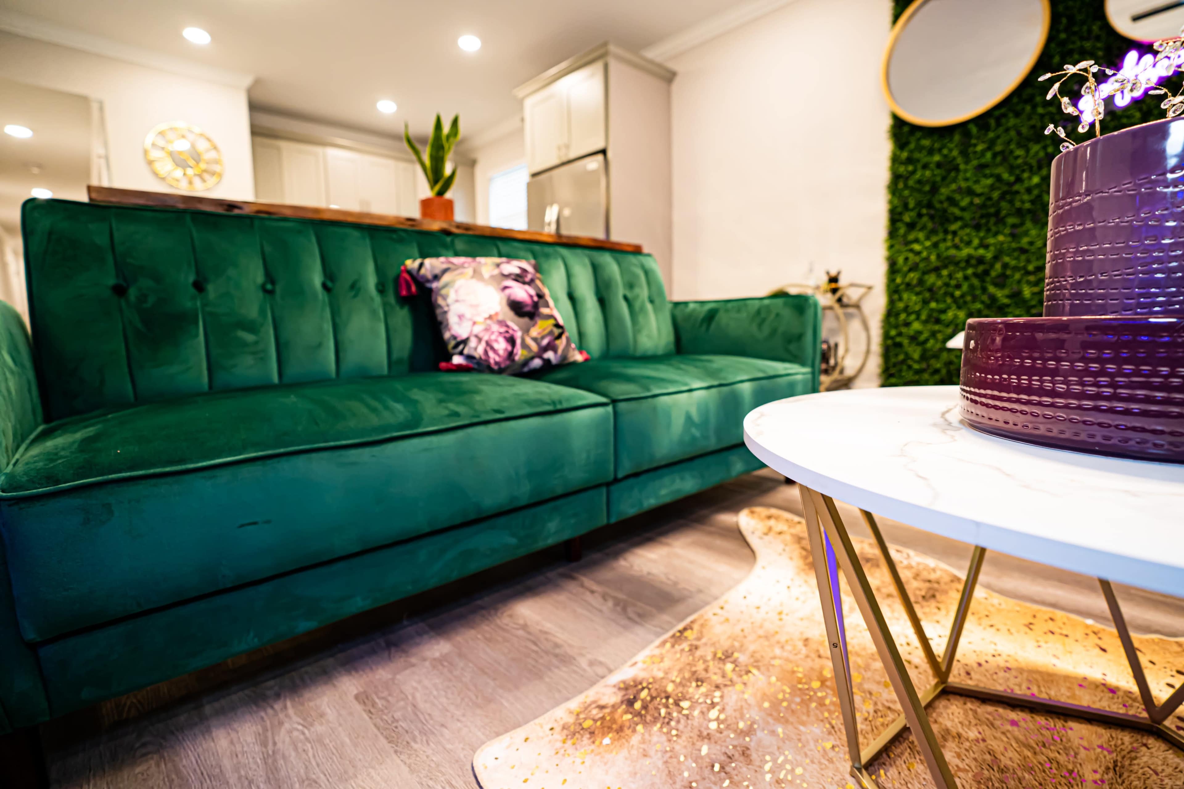 The image shows a green velvet sofa with a decorative pillow, positioned next to a round marble table on a gold frame, in a modern living space featuring a green wall and overhead lighting.