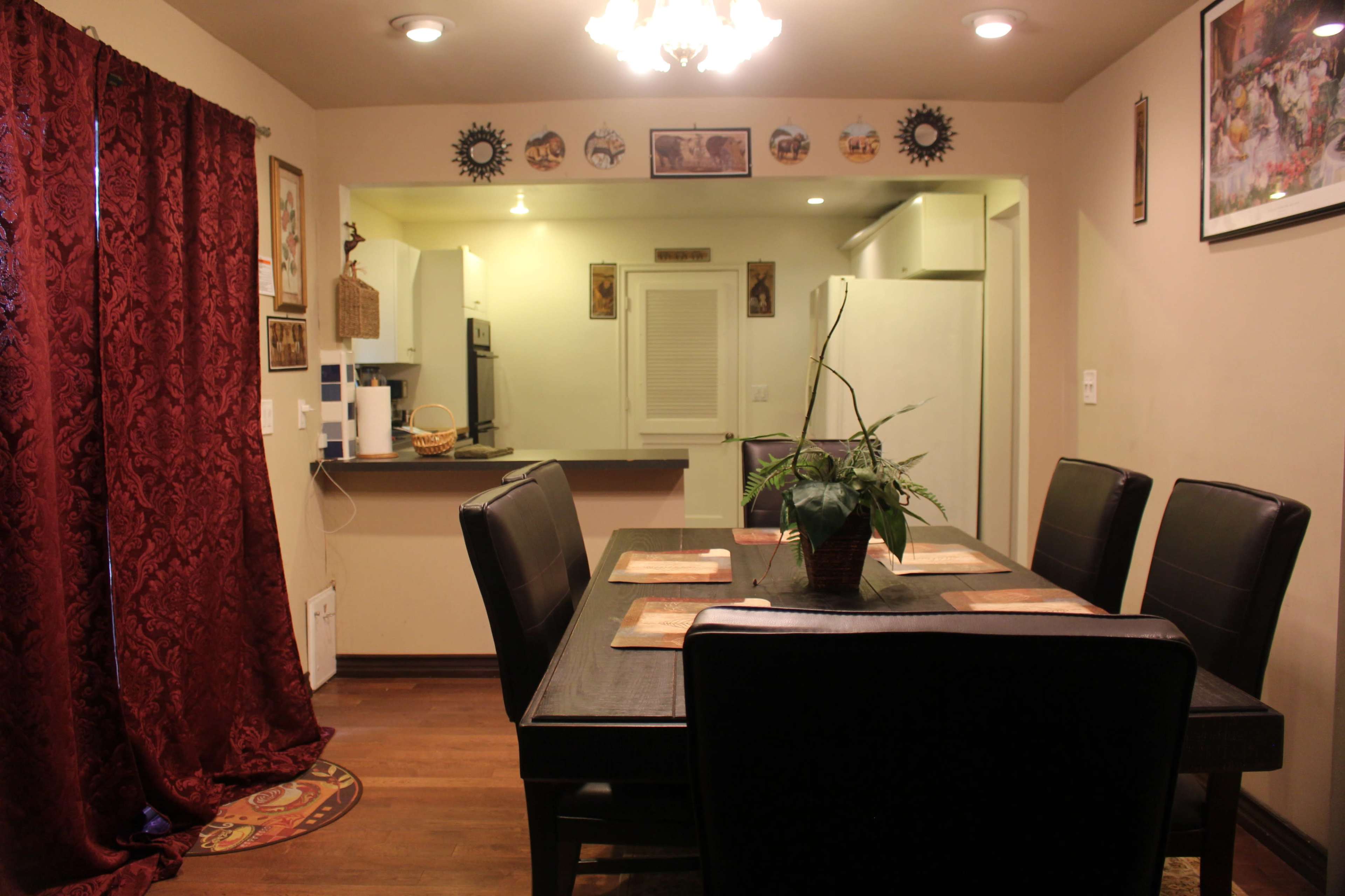 A dining area features a large table with black chairs, surrounded by decorative curtains and wall art, leading to an adjacent kitchen space.